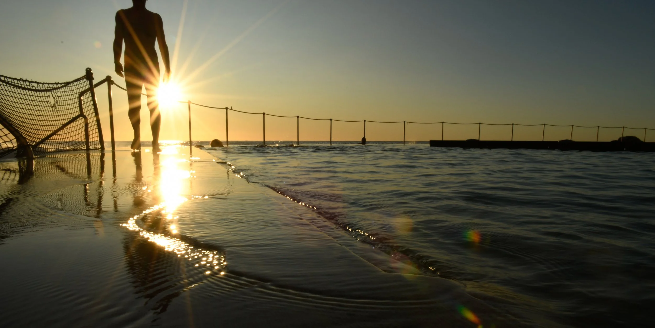 Silhouette of a person walking near the sea at sunset with the sun shining behind them, casting reflections on the water, and a safety net along the edge of a swimming pool.