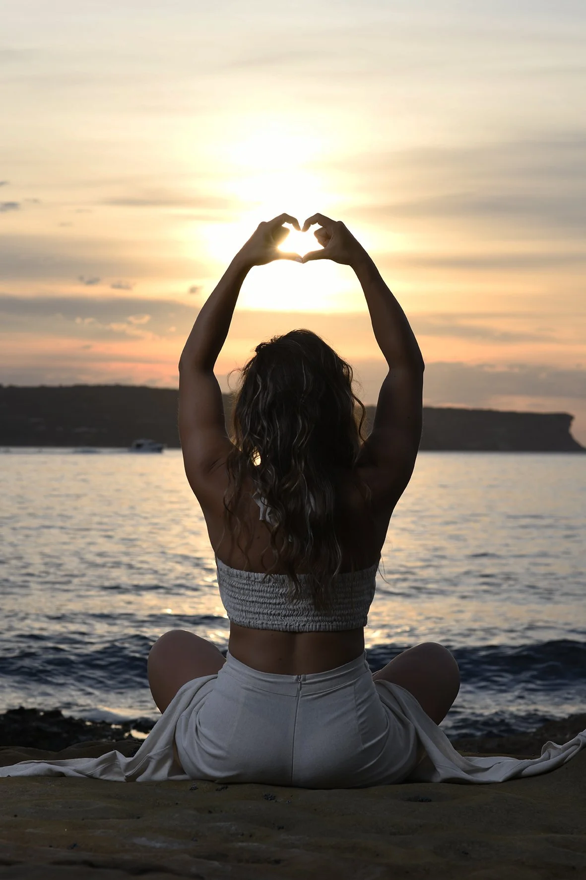 A woman sitting cross-legged at the beach during sunset, forming a heart shape with her hands around the sun.