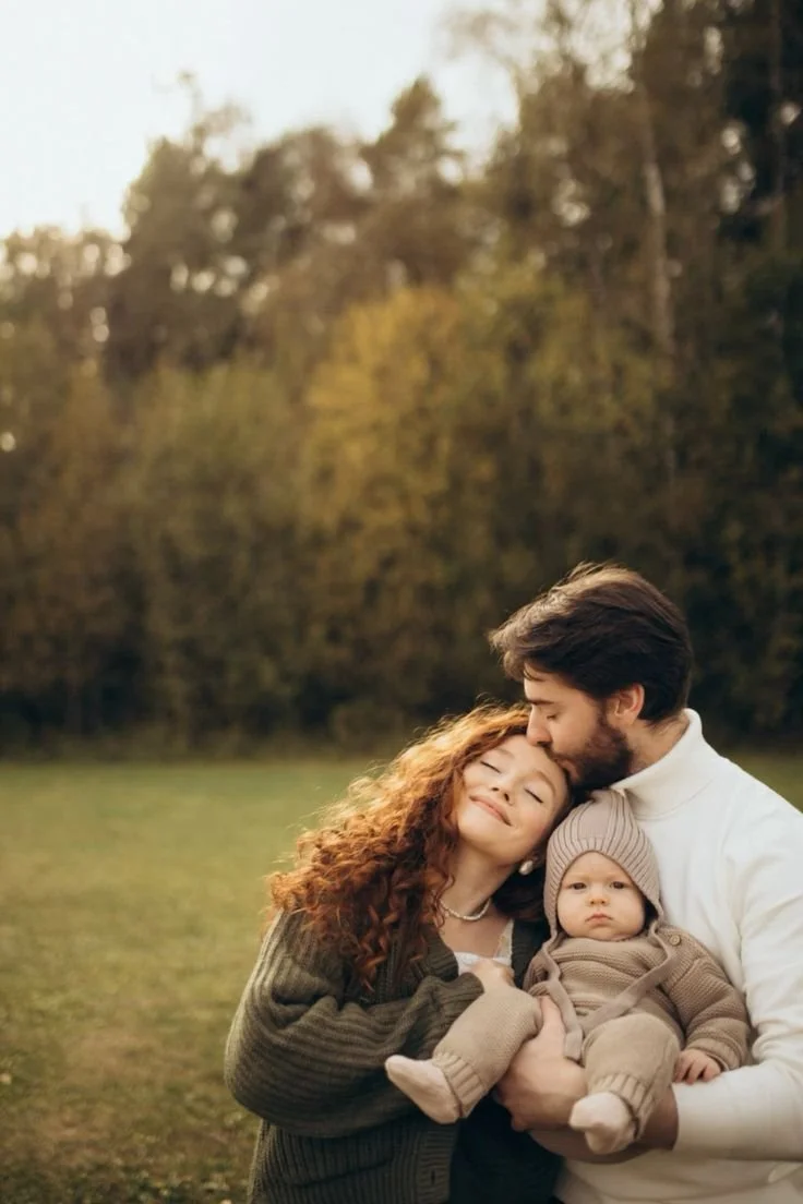 A family of three, a woman, man, and a baby, standing outdoors in a grassy field with trees in the background during sunset. The man is kissing the woman on the forehead while holding the baby. The woman has curly hair, and the baby is wearing a beige outfit and hat.