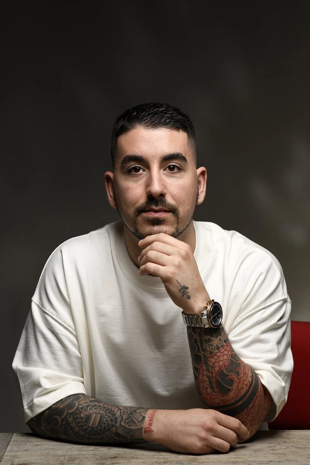 A young man with short dark hair, a mustache, and a goatee, wearing a white t-shirt, resting his chin on his hand. He has tattoos on his left arm and wrist, and a watch on his left wrist. The background is dark and neutral.