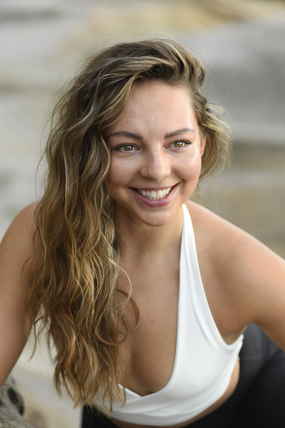 Close-up of a smiling woman with long wavy brown hair, wearing a white tank top, outdoors near water.