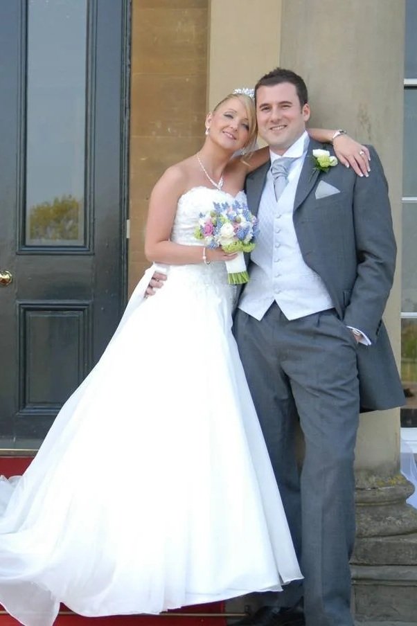 A bride and groom standing together outside a building, smiling. The bride is in a white wedding dress holding a bouquet, and the groom is in a gray suit with a light blue vest and tie, with his hands in his pockets.