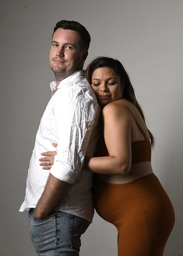 A man and woman stand close together, with the woman hugging and resting her head on the man's shoulder. The man has short dark hair, is wearing a white button-up shirt with rolled sleeves, and jeans. The woman has long dark hair, is wearing a brown crop top and matching high-waisted pants, and appears to be content and relaxed with her eyes closed.