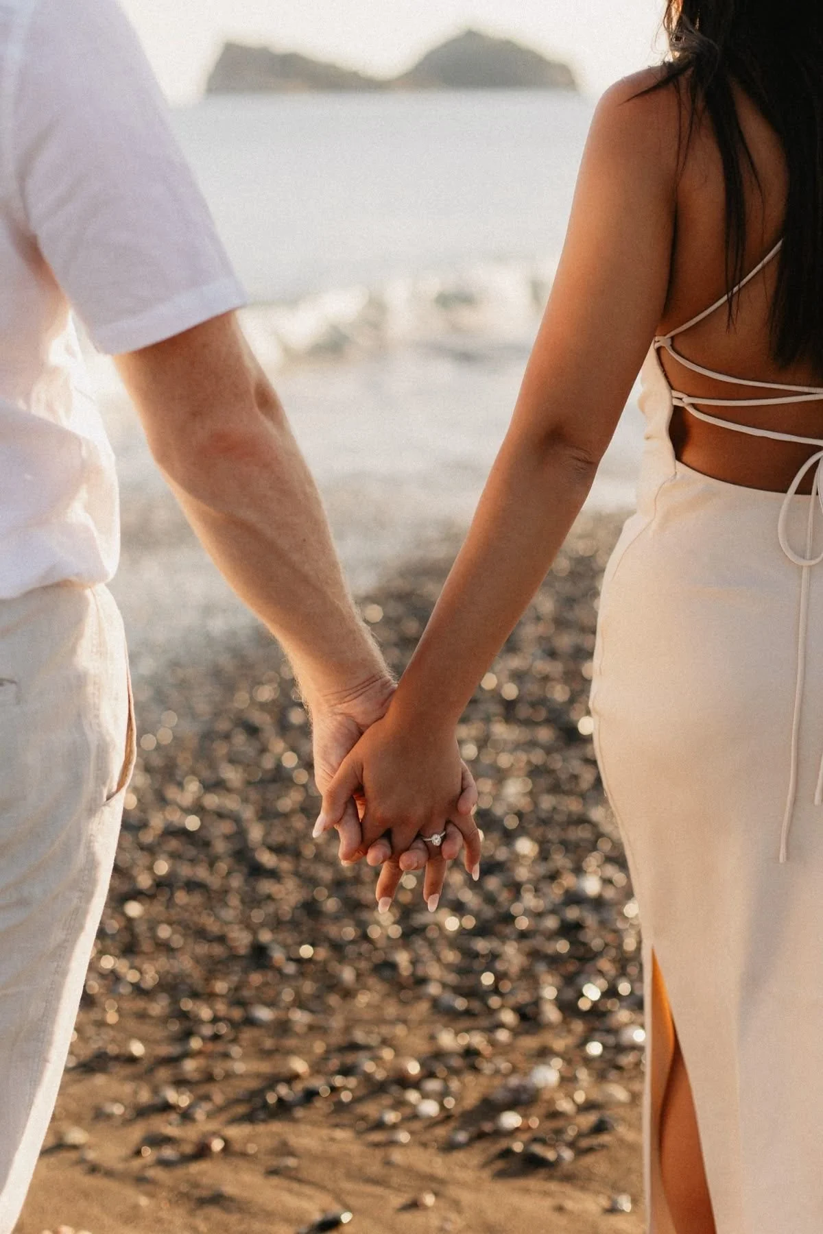 A couple holding hands on a beach at sunset, with the ocean and distant mountains in the background.