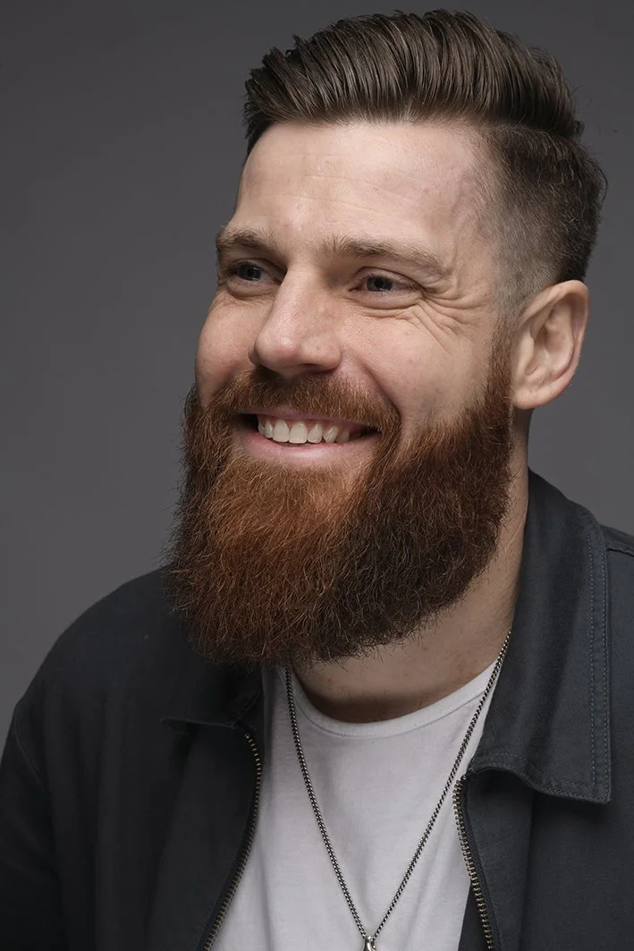 A smiling man with a full, reddish-brown beard and styled hair, wearing a black jacket over a white shirt and a necklace, against a gray background.