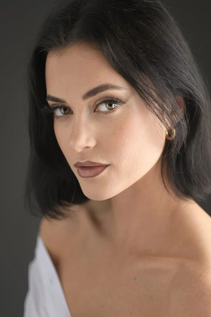 Close-up portrait of a woman with black hair, green eyes, and makeup, wearing a white top and gold hoop earrings, against a dark background.