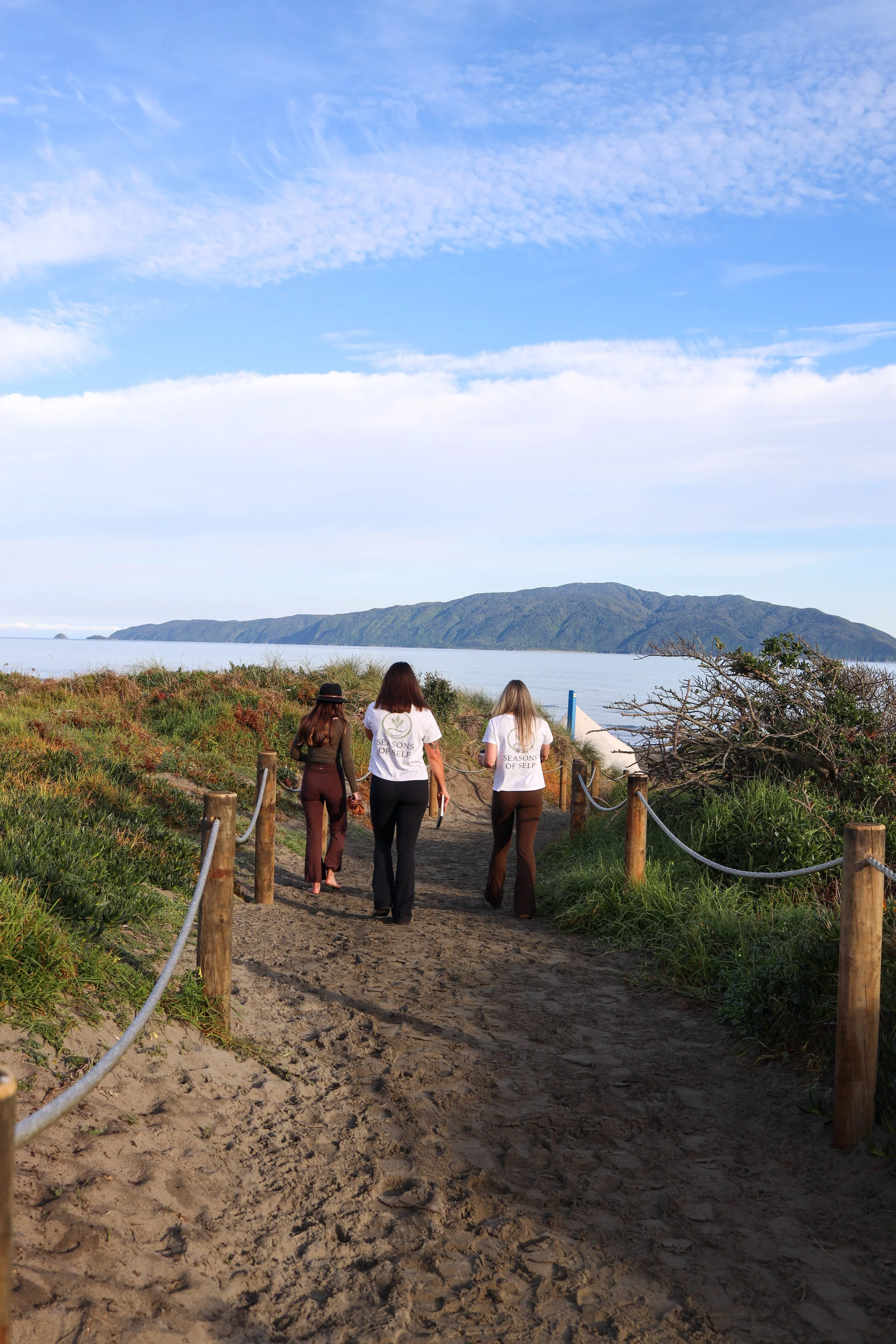 Three people walking along a sandy path bordered by wooden posts and rope, with a body of water and a mountain in the background under a partly cloudy sky.