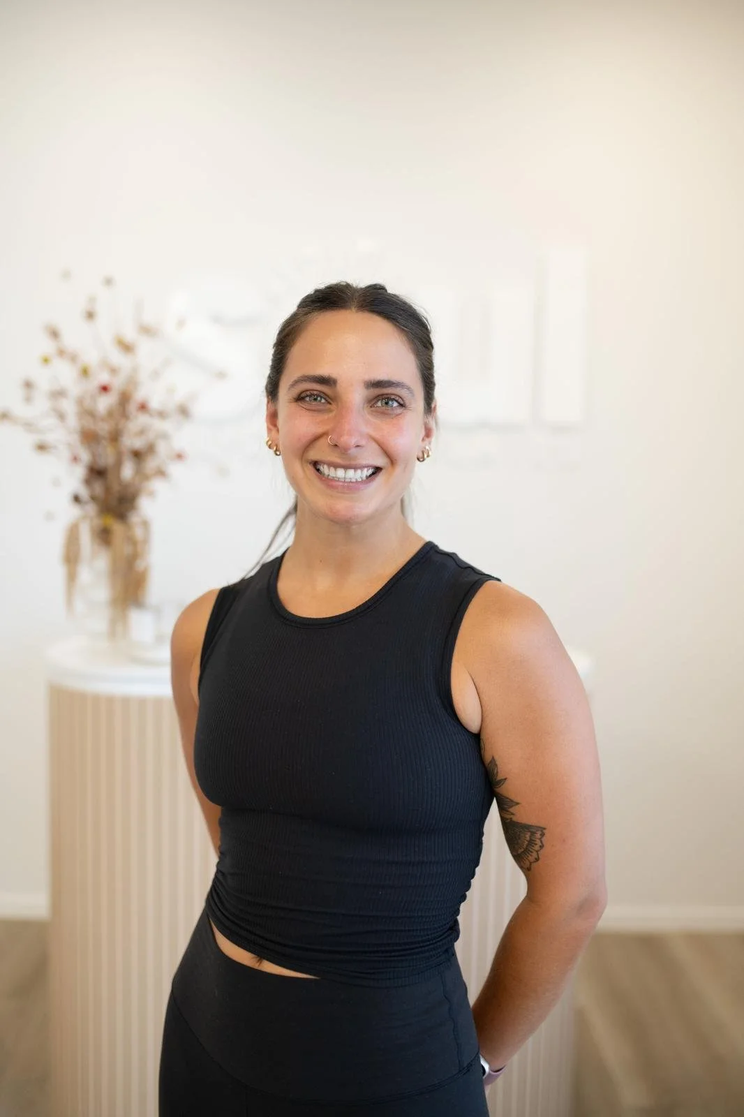 A woman in a black sleeveless top smiling at the camera, standing indoors with a blurred background of a white wall and a table with a vase of dried flowers.