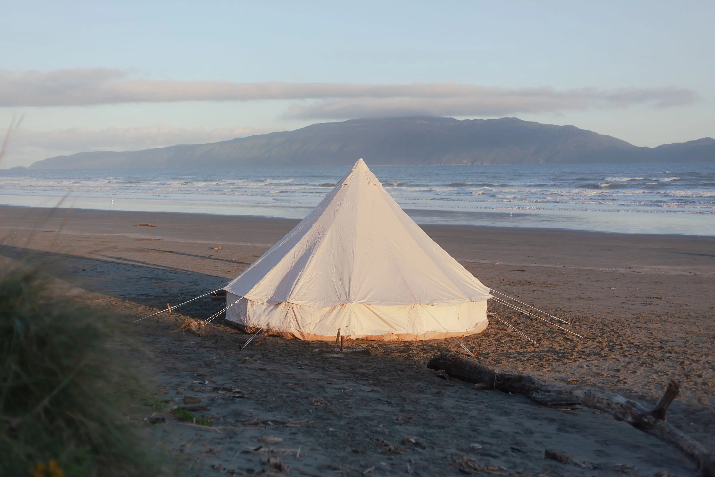 A white tent set up on a sandy beach with ocean waves and a mountainous island in the background under a partly cloudy sky.