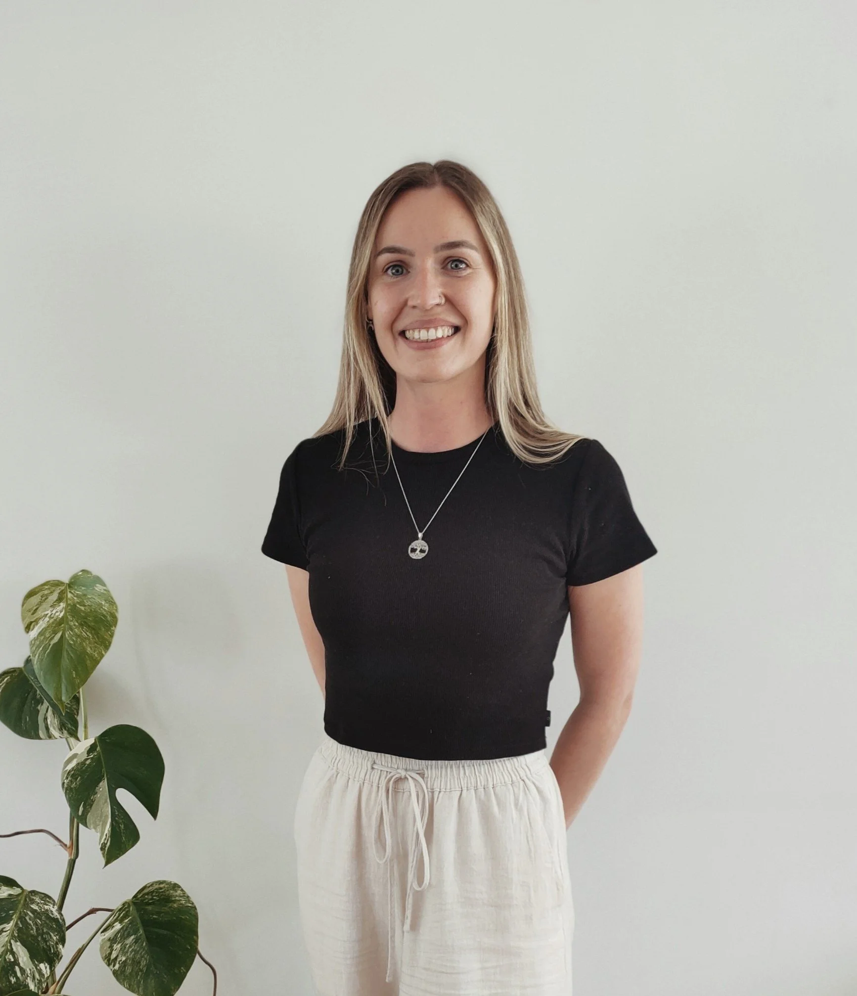 A young woman with long blonde hair smiling while standing against a plain light-colored wall. She is wearing a black t-shirt, light-colored linen pants with a drawstring, a silver necklace with a skull pendant, and small earrings. There is a green variegated pothos plant in the lower left corner of the image.