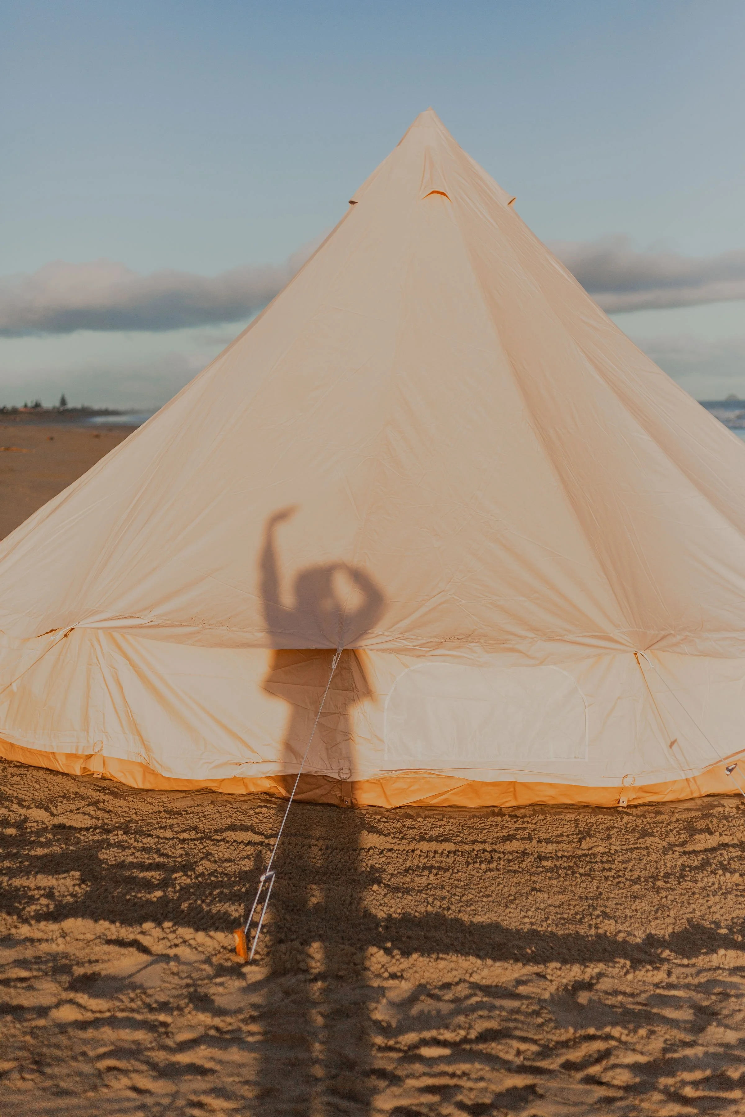 A cream-colored tent set up on a sandy beach with a partly cloudy sky and the ocean in the background. The tent is illuminated by sunlight, and shadows of a person and the tent's support ropes are visible on the sand.