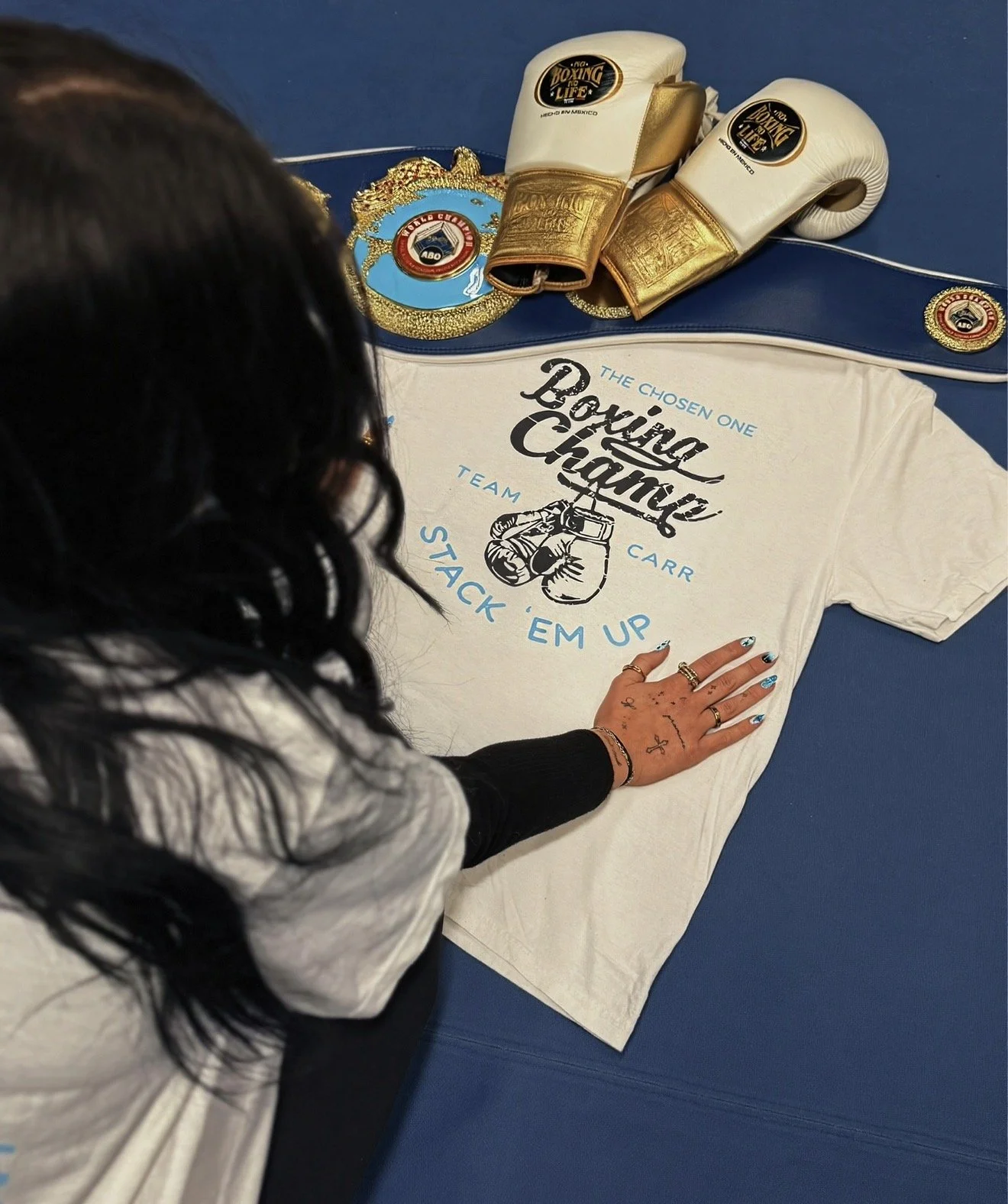 Boxing gloves, a championship belt, and a t-shirt with boxing-related text are on a blue floor. A person with tattoos on their hand and long dark hair is touching the t-shirt.