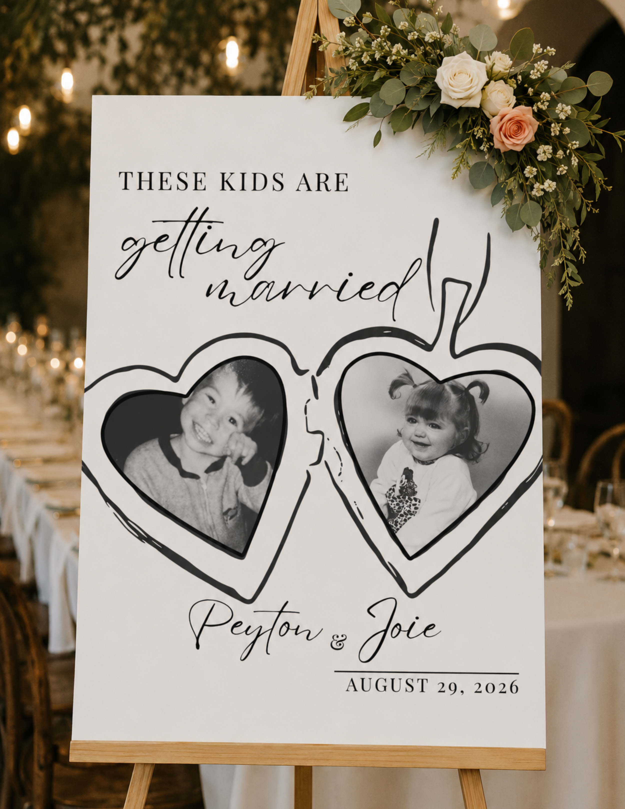 Wedding sign with black and white photographs of children inside heart-shaped outlines, decorated with a floral arrangement of white and pink roses and green leaves, displaying the names Peyton and Joie and the date August 29, 2026.