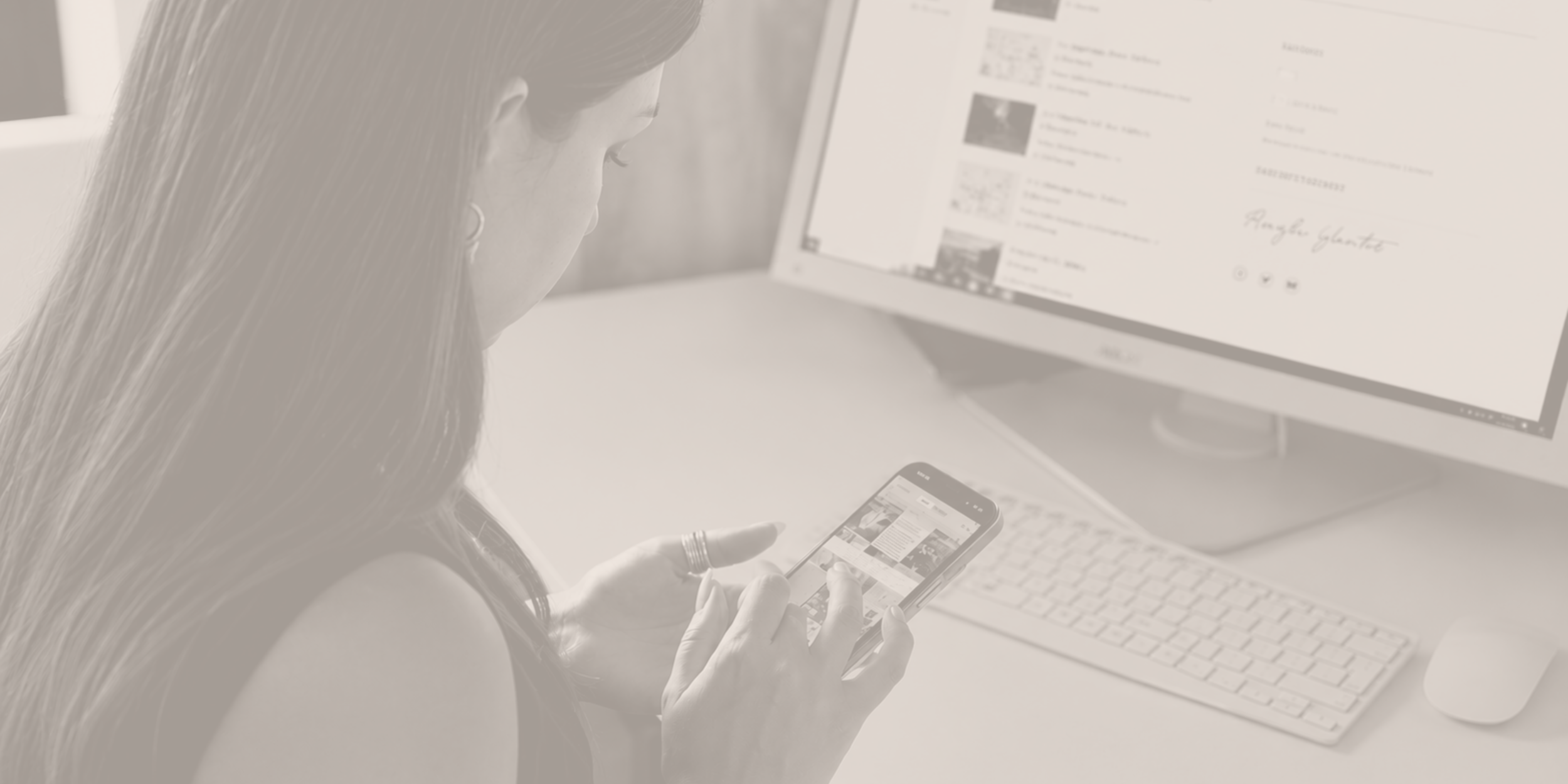 Woman sitting at desk looking at her smartphone with a computer monitor showing a social media feed.