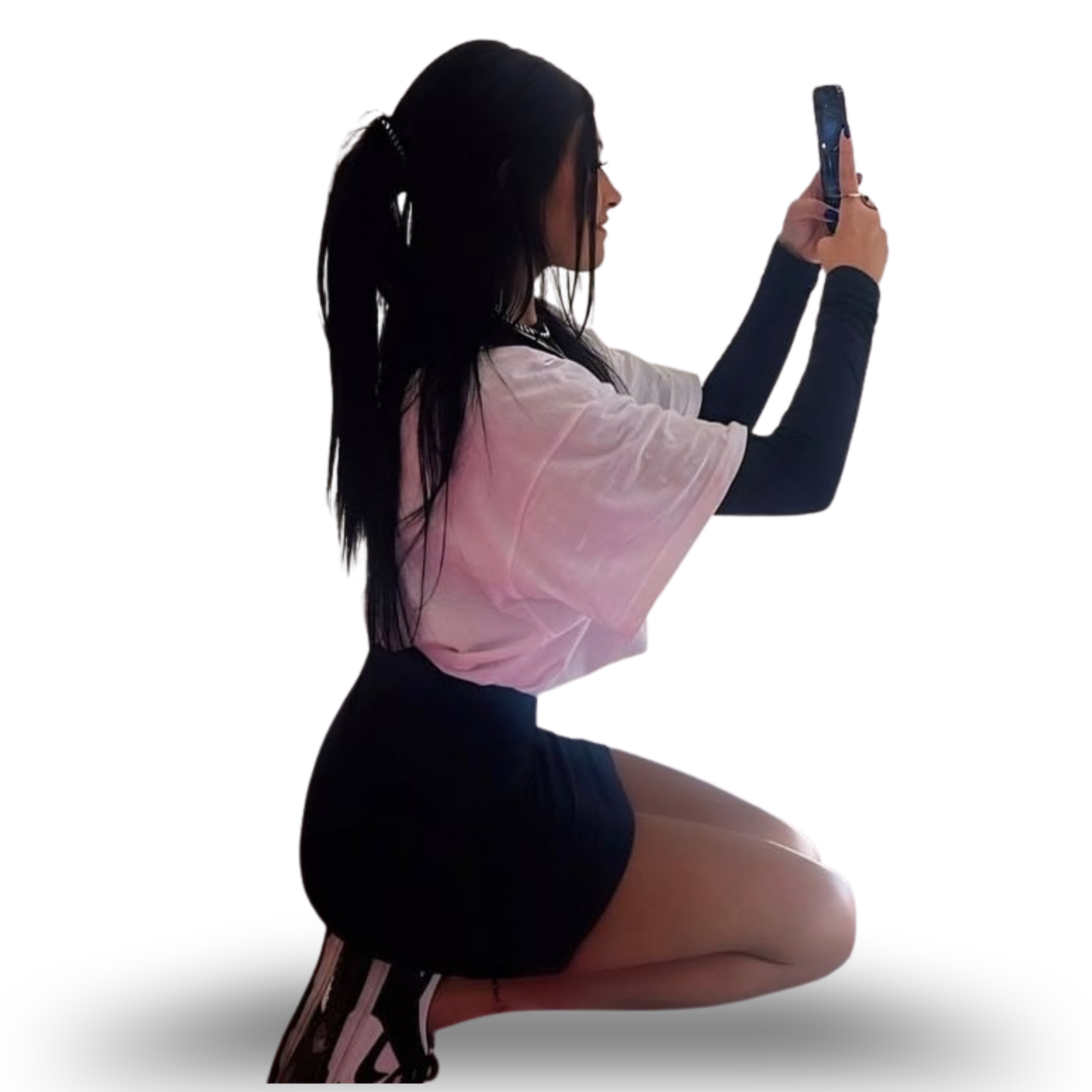 A woman with long dark hair in a ponytail, wearing a black top and jeans, is taking a selfie with her smartphone against a black background.