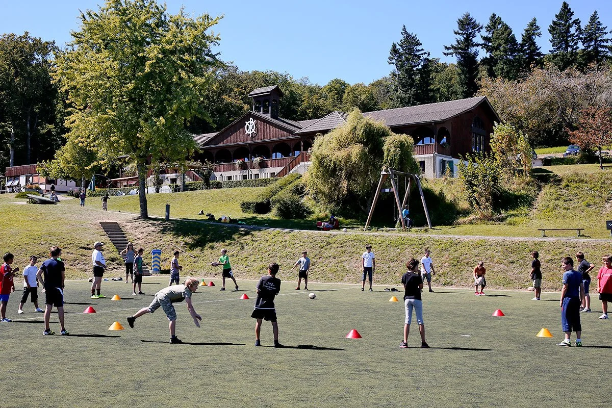 Groupe d'enfants jouant au football sur un terrain en plein air, avec un bâtiment en bois et des arbres en arrière-plan.