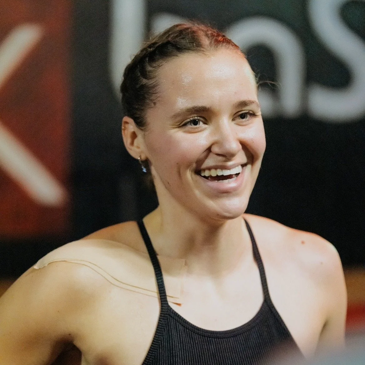 Une jeune femme souriante avec des cheveux attachés, portant un haut de sport noir, dans une salle de sport.