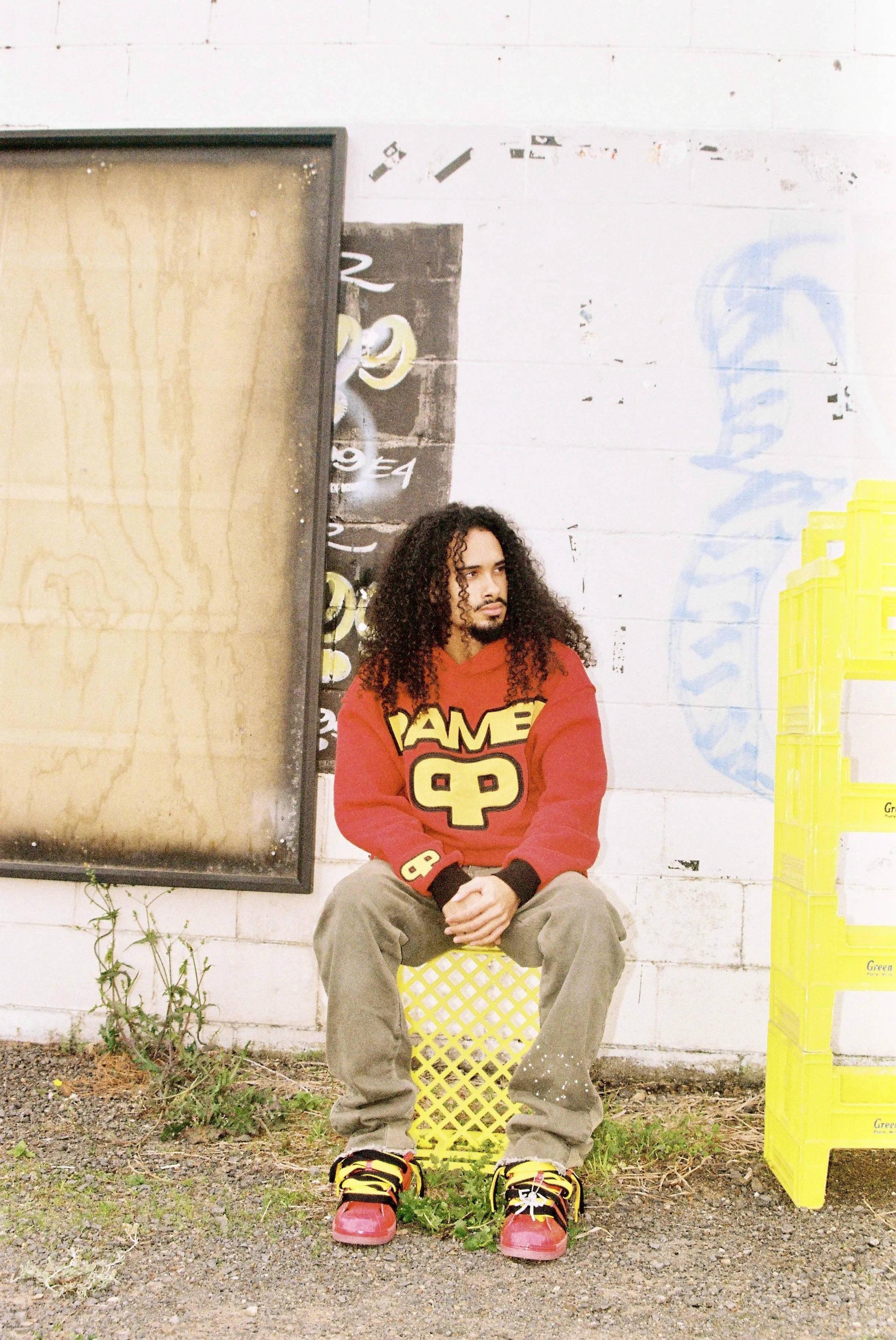A man with long curly hair wearing a red sweatshirt with yellow and black graphics sitting on a yellow lattice-patterned crate outside on gravel and dirt ground, next to yellow crates and a white wall with graffiti.