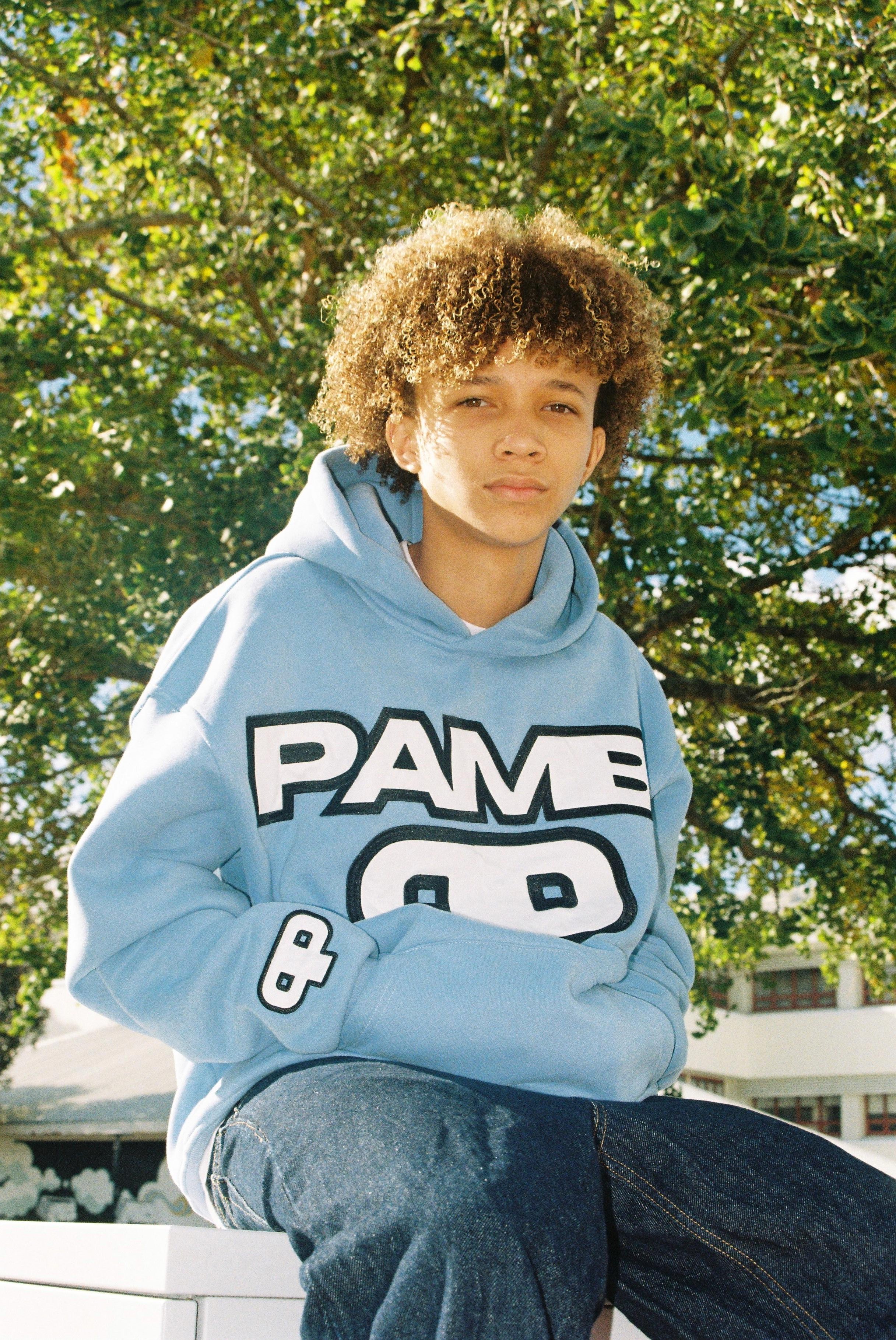 A young person with curly hair wearing a light blue hoodie and dark jeans, sitting outdoors on a sunny day with green trees in the background.