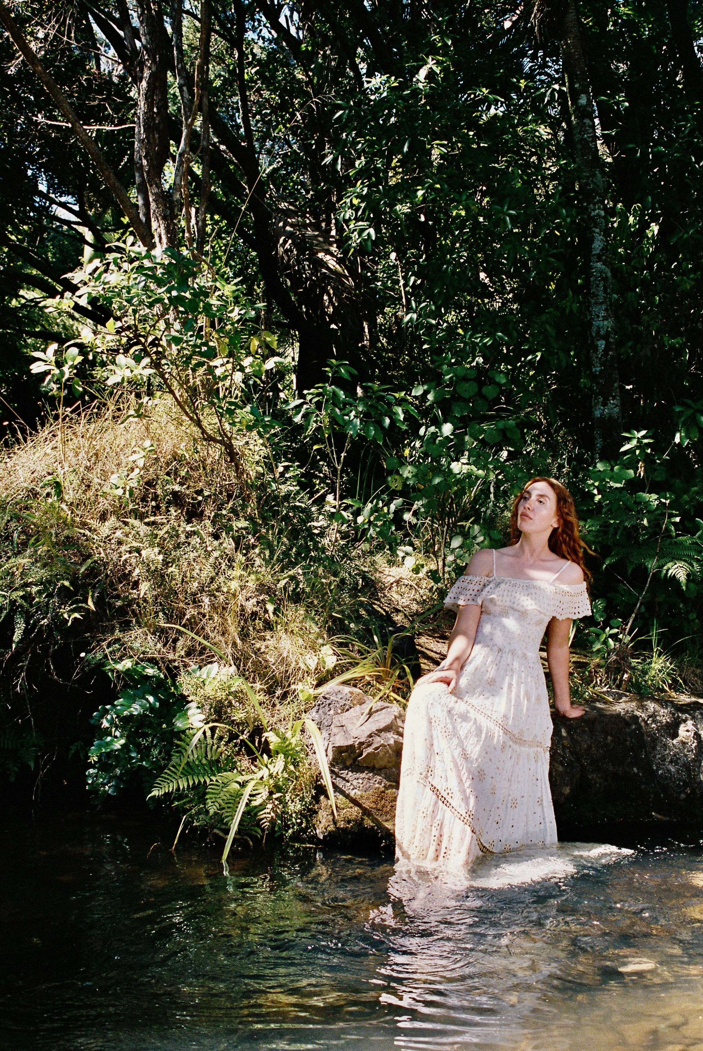 A woman in a white dress with lace details sitting on a rock by a stream in a forest, surrounded by lush green foliage and sunlight filtering through the trees.