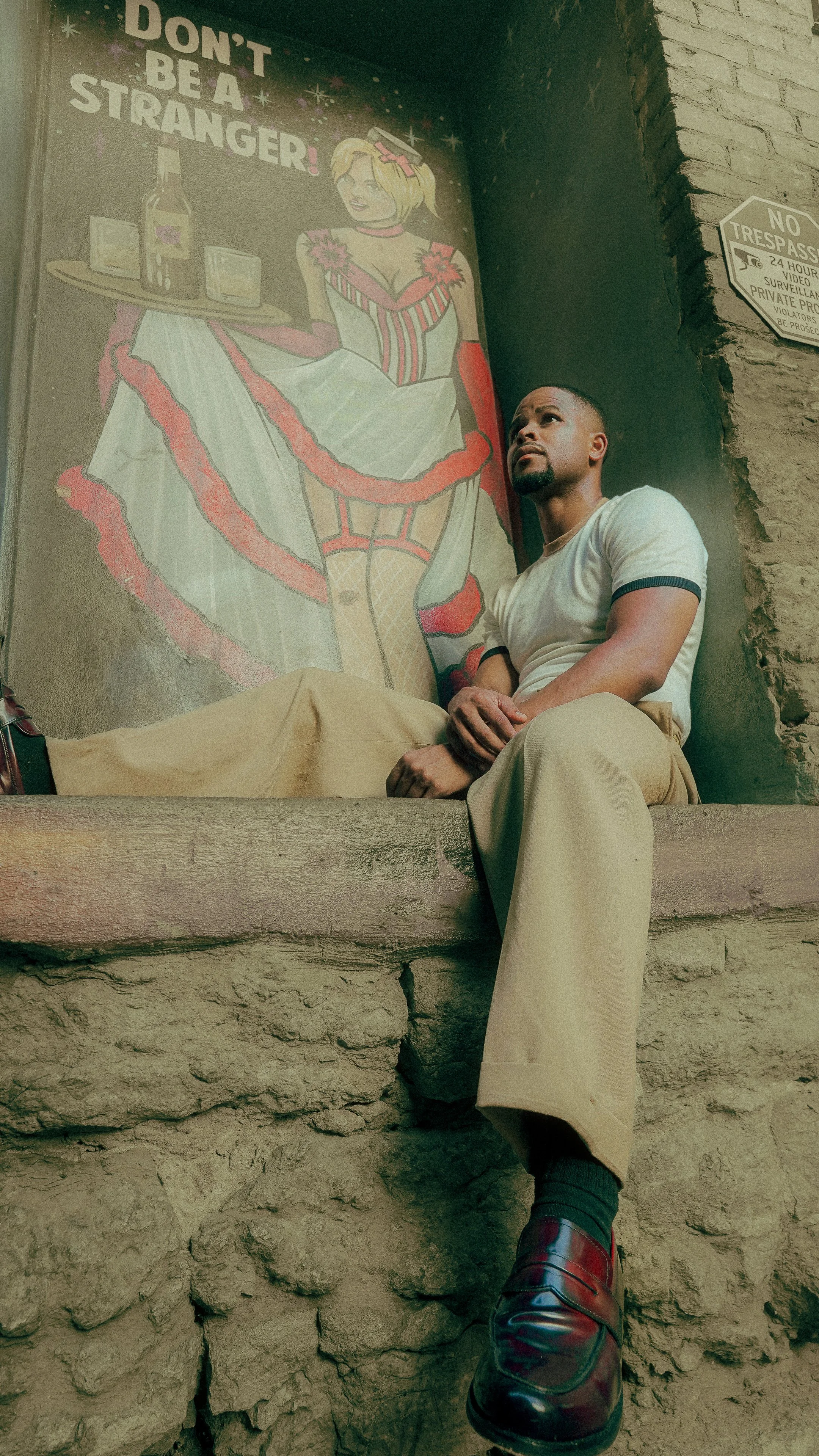 A man sitting on a ledge in front of a painted wall. The painting depicts a cartoon waitress holding a tray with bottles and glasses, wearing a revealing outfit, with the phrase "Don't be a stranger!" written above.