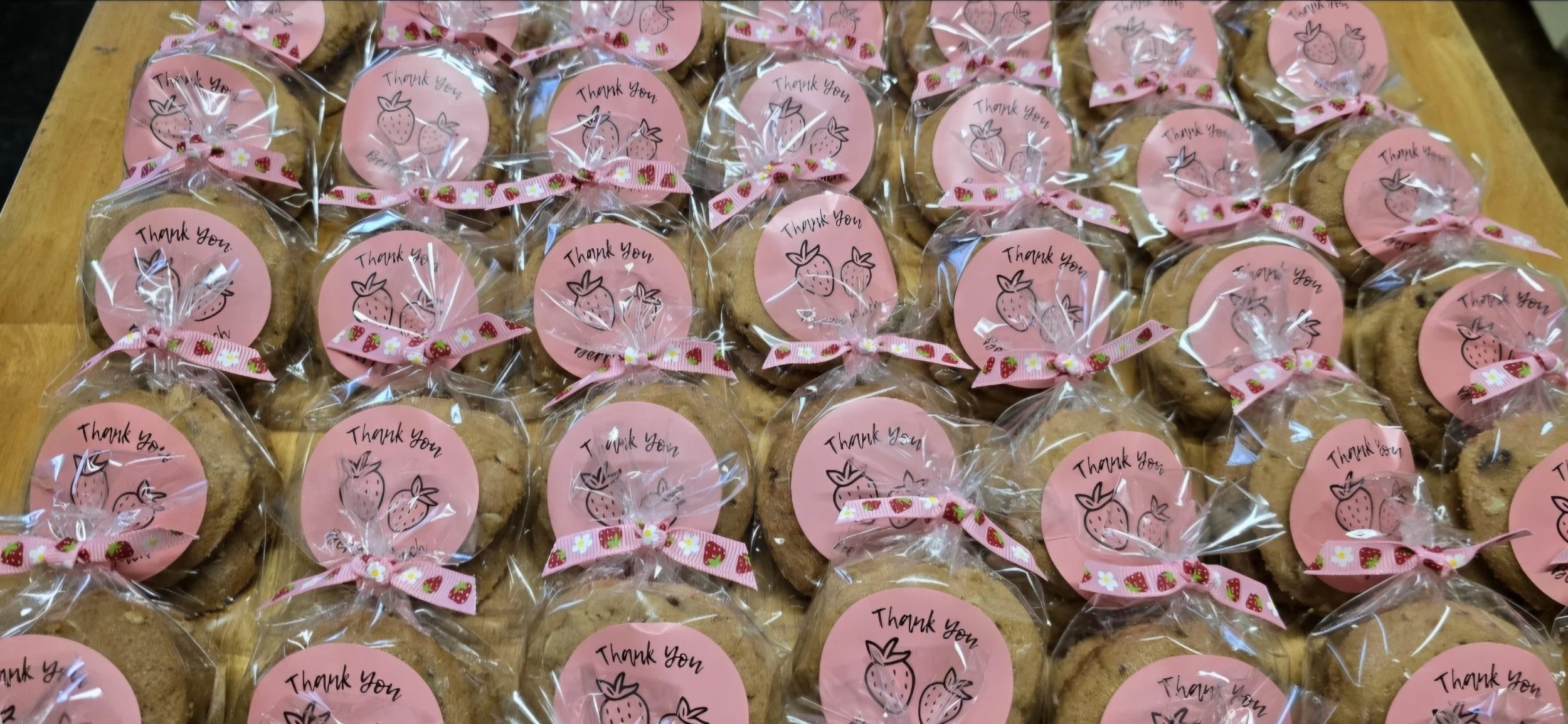A collection of individually wrapped cookies decorated with pink circular tags that read 'Thank you' and feature strawberry illustrations, tied with pink ribbon with strawberry patterns, on a wooden surface.