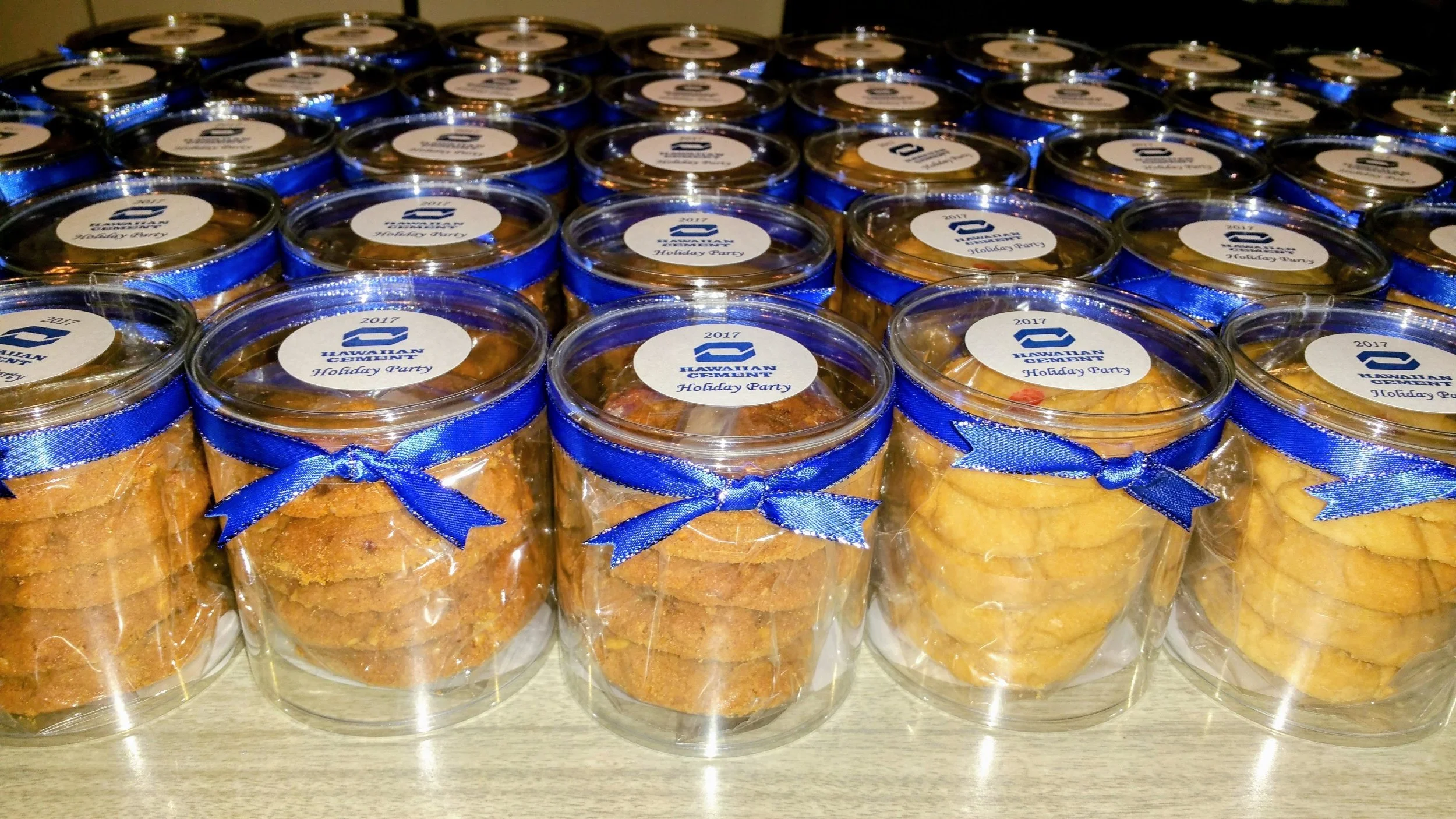 Multiple clear plastic jars filled with cookies, each tied with a blue ribbon and labeled with '2017 Hawaiian Government Holiday Party'.