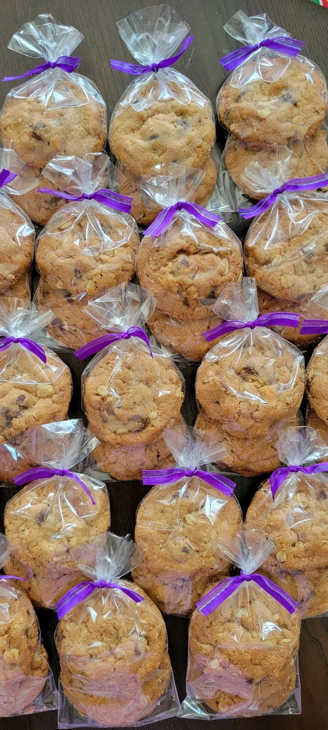 Assorted cookies wrapped in clear plastic bags tied with purple ribbons, laid out on a wooden surface.