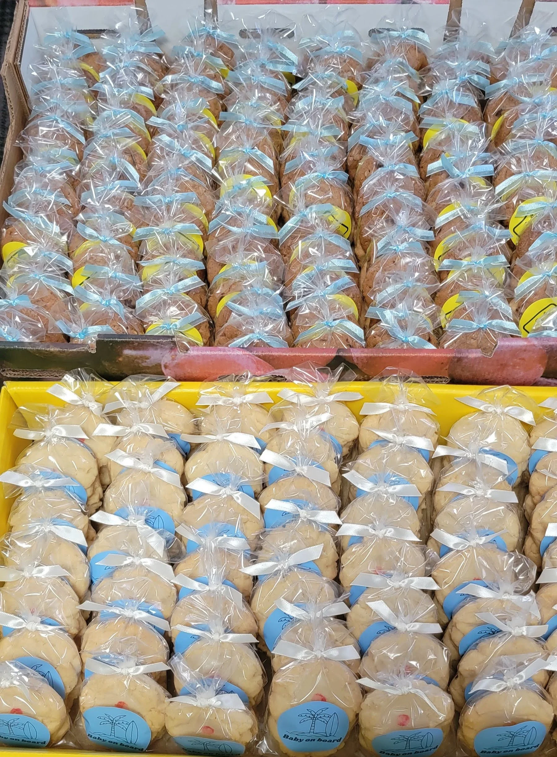 Multiple cookies in clear plastic bags tied with blue and white ribbons, laid out in trays with a label reading "Baby on board," possibly for a baby shower or party.
