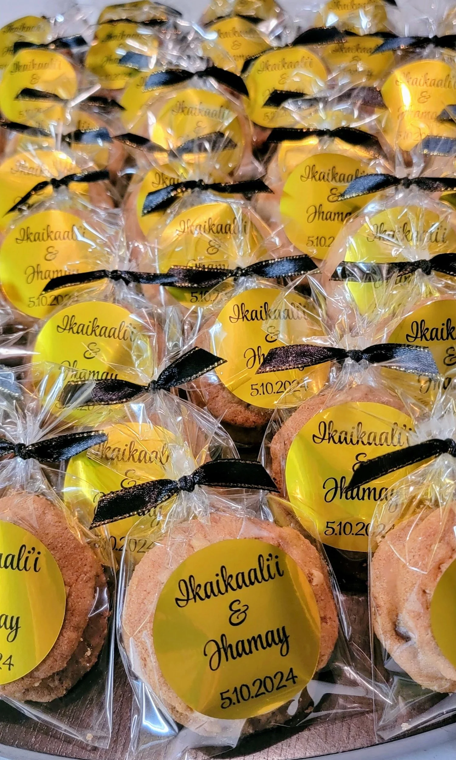 Decorative cookies wrapped in clear plastic with black ribbons, each featuring a yellow label with the names 'Ikaikaali & Jhamay' and the date 5.10.2024.