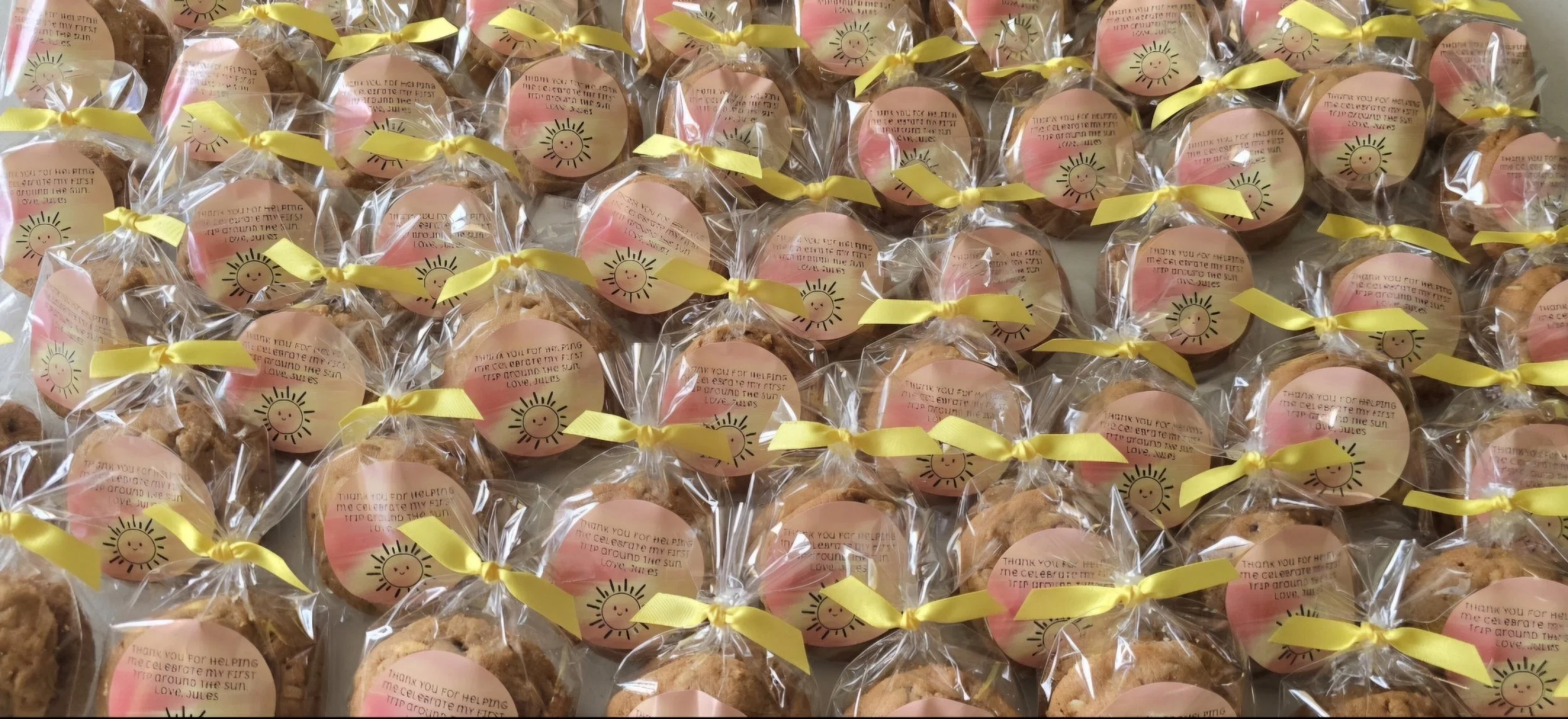 A large pile of individually wrapped cookies, each with a pink sticker featuring a smiling sun drawing and a thank you message, tied with yellow ribbons.