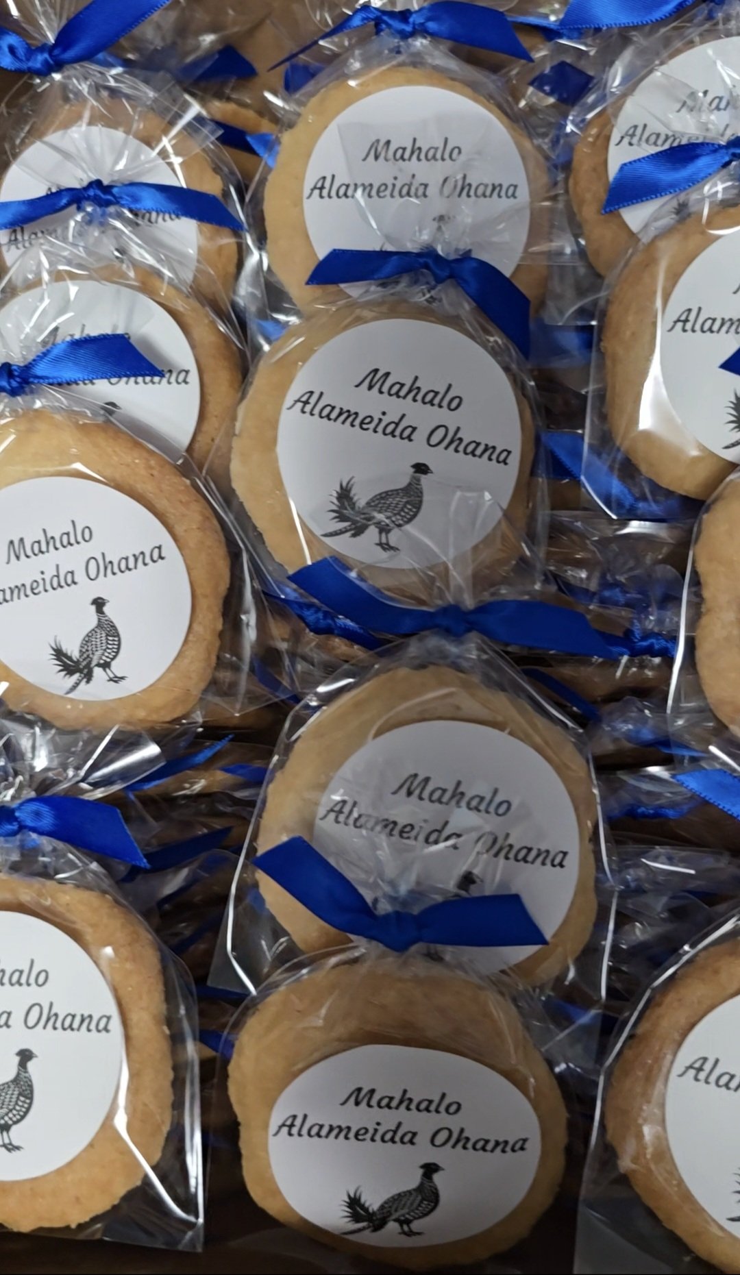 Cookies wrapped in clear plastic with a blue ribbon, each with a white circle label that says 'Mahalo Almeida Ohana' with a small illustration of a bird.