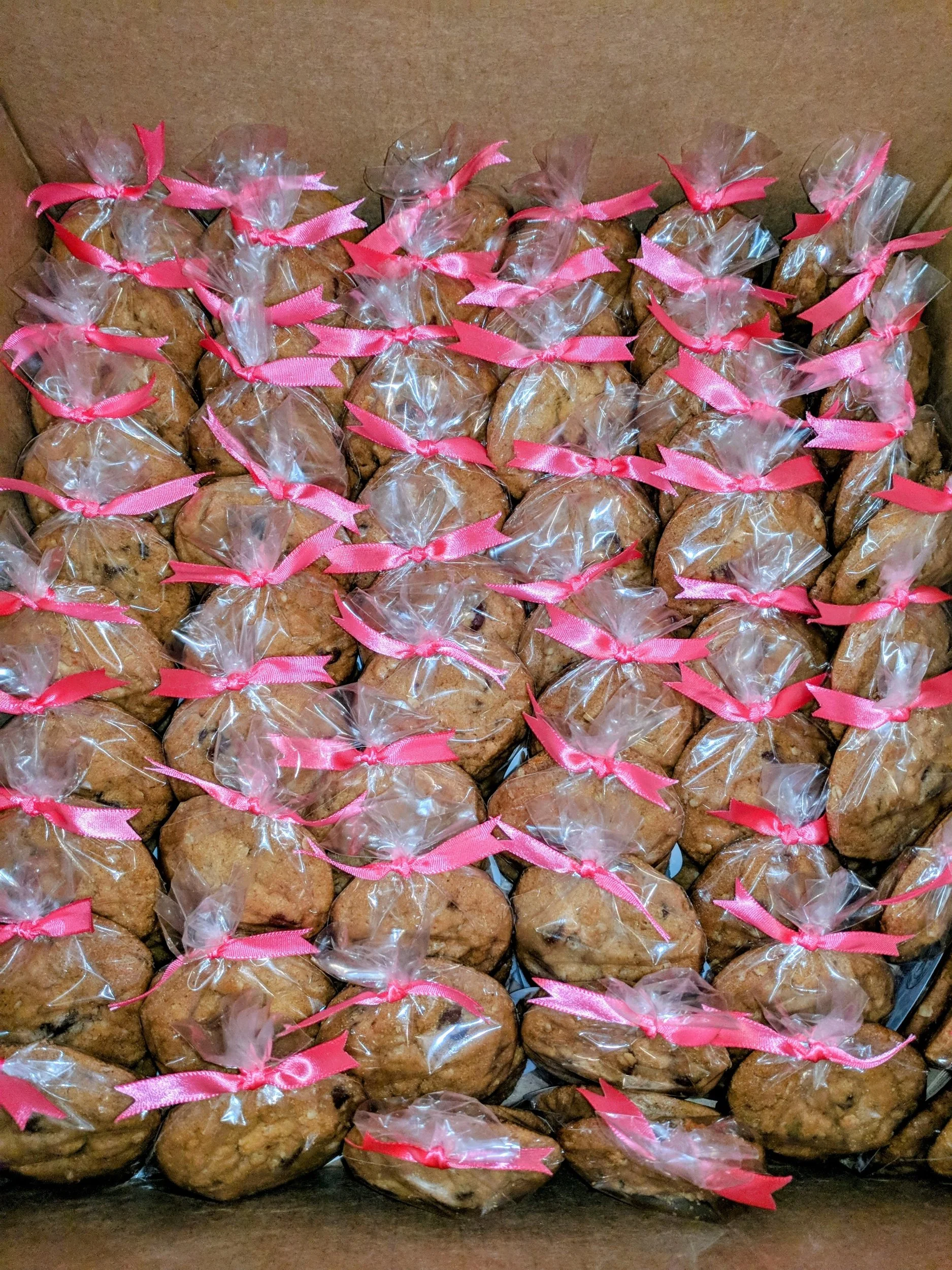 Multiple baked goods wrapped in clear plastic bags tied with pink ribbons inside a cardboard box.