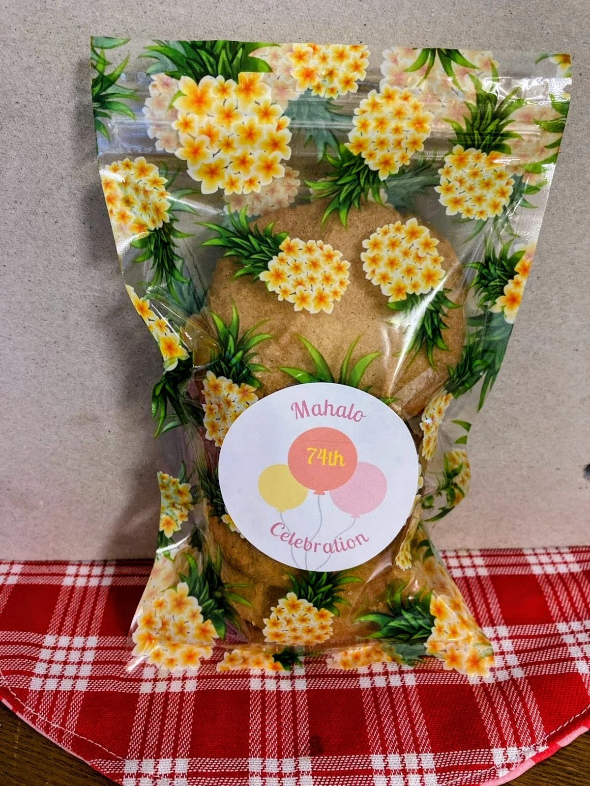 Decorative plastic bag with yellow and orange flowers and green leaves, containing cookies, with a birthday label reading 'Mahalo 74th Celebration' placed on a red and white checkered cloth.