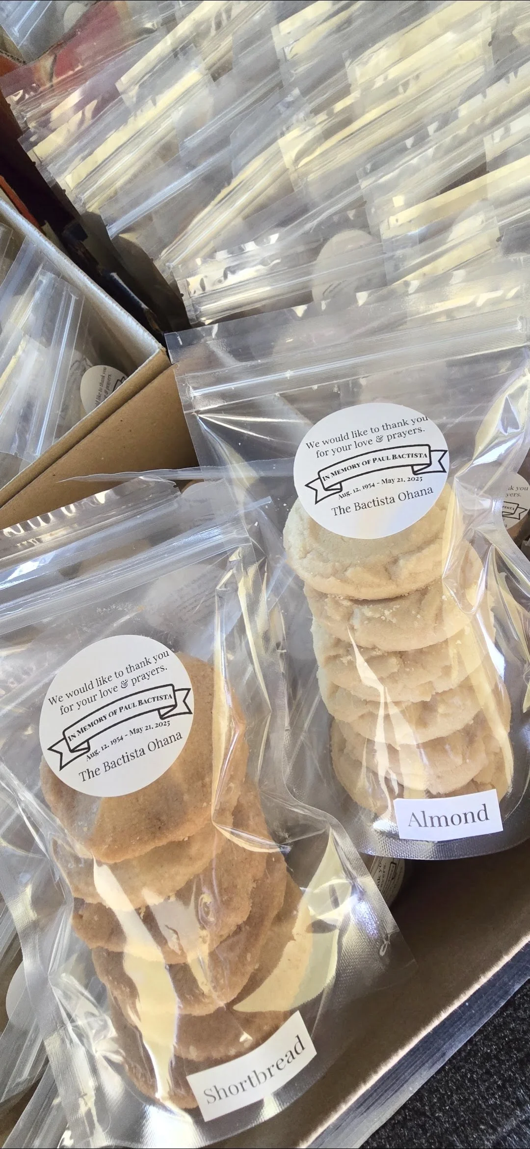 Clear plastic bags of almond shortbread cookies with memorial labels and a small white sticker labeled 'Almond' on one of the bags, displayed on a metallic surface.