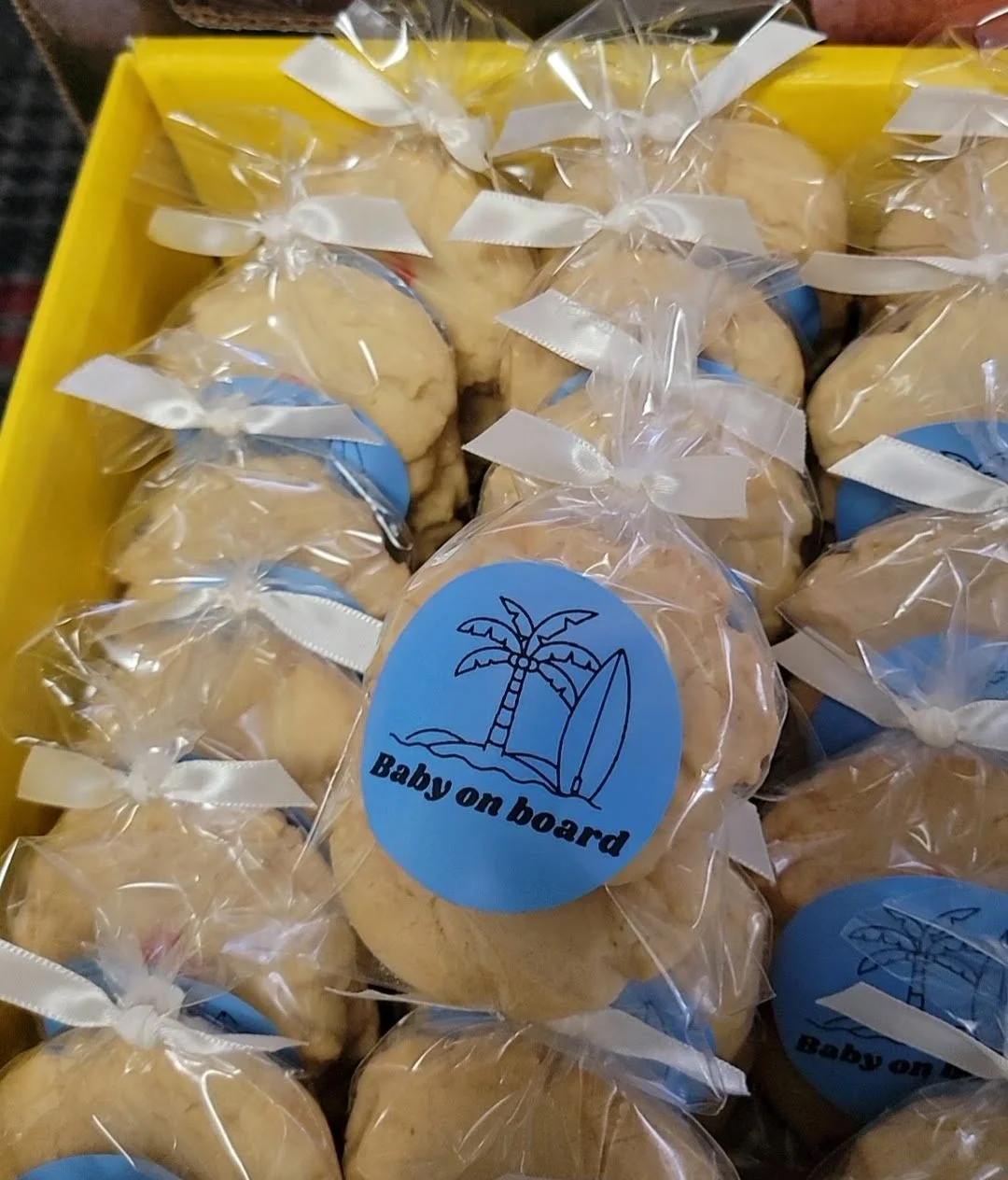 Cookies wrapped in clear plastic bags with white ribbons, in a yellow box, labeled "Baby on board" with a blue sticker featuring a palm tree and a surfboard.