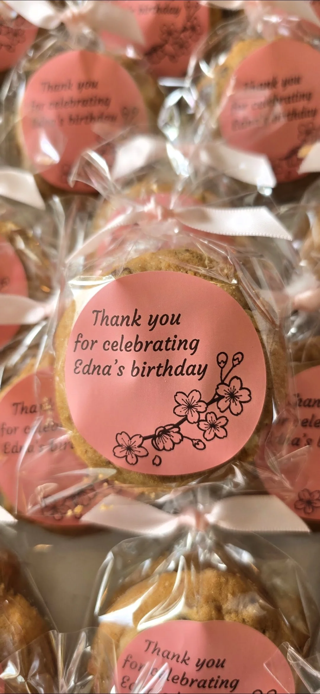 Individual cookies wrapped in clear plastic bags with pink circular labels that say "Thank you for celebrating Edna's birthday" with a cherry blossom illustration.