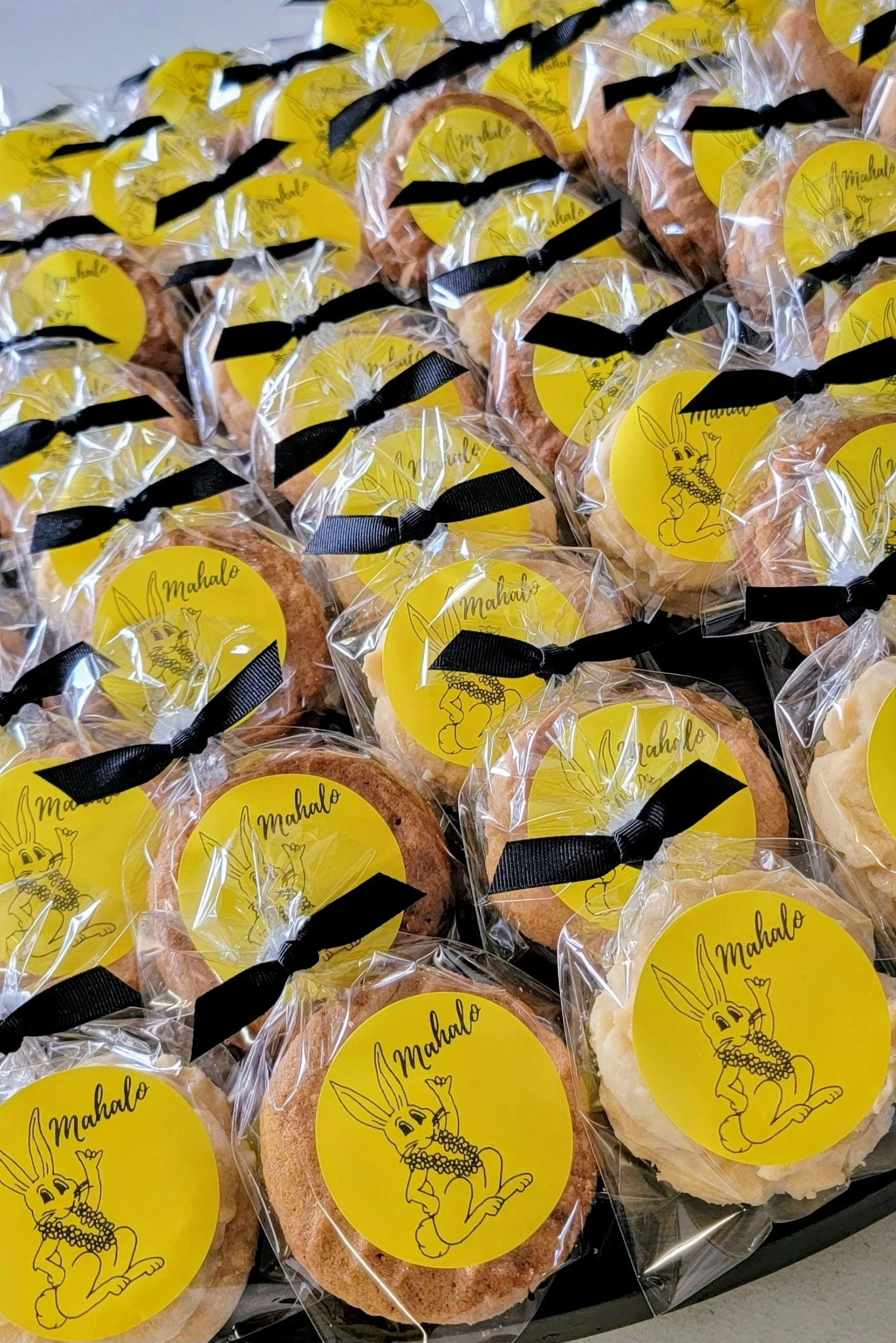 Several decorated cookies individually wrapped in clear plastic with yellow labels featuring a cartoon rabbit character and the word 'Mahalo,' tied with black ribbons.