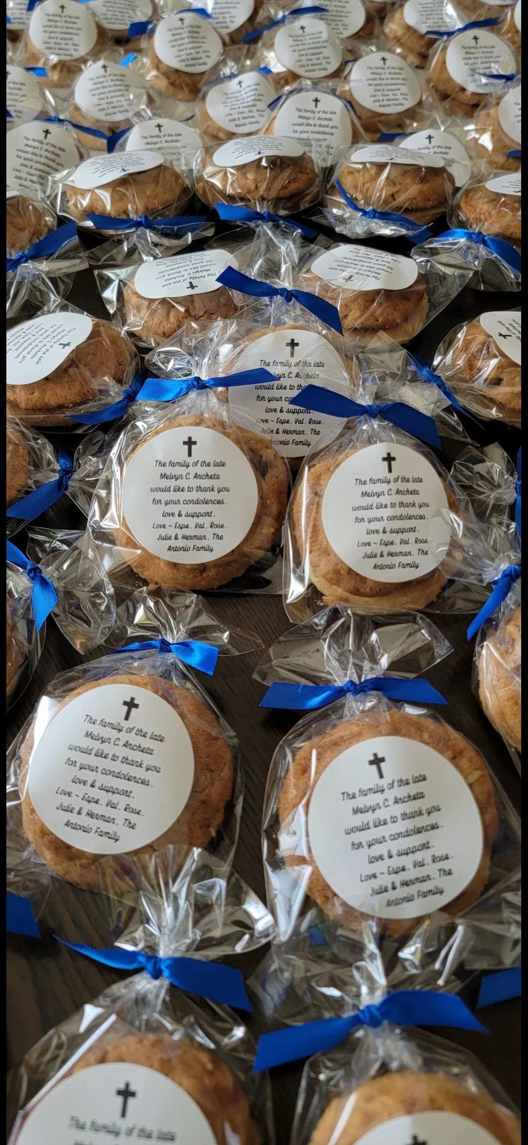 Multiple packaged cookies wrapped in clear plastic with blue ribbons, each with a white circular label featuring a cross and a message of condolence from the family of the late Mellyn C. Ancheta.