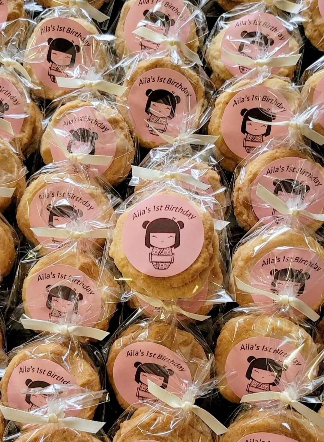 Cookies wrapped in clear plastic with a pink sticker featuring a cartoon girl in traditional Japanese attire, celebrating Aila's 1st birthday.
