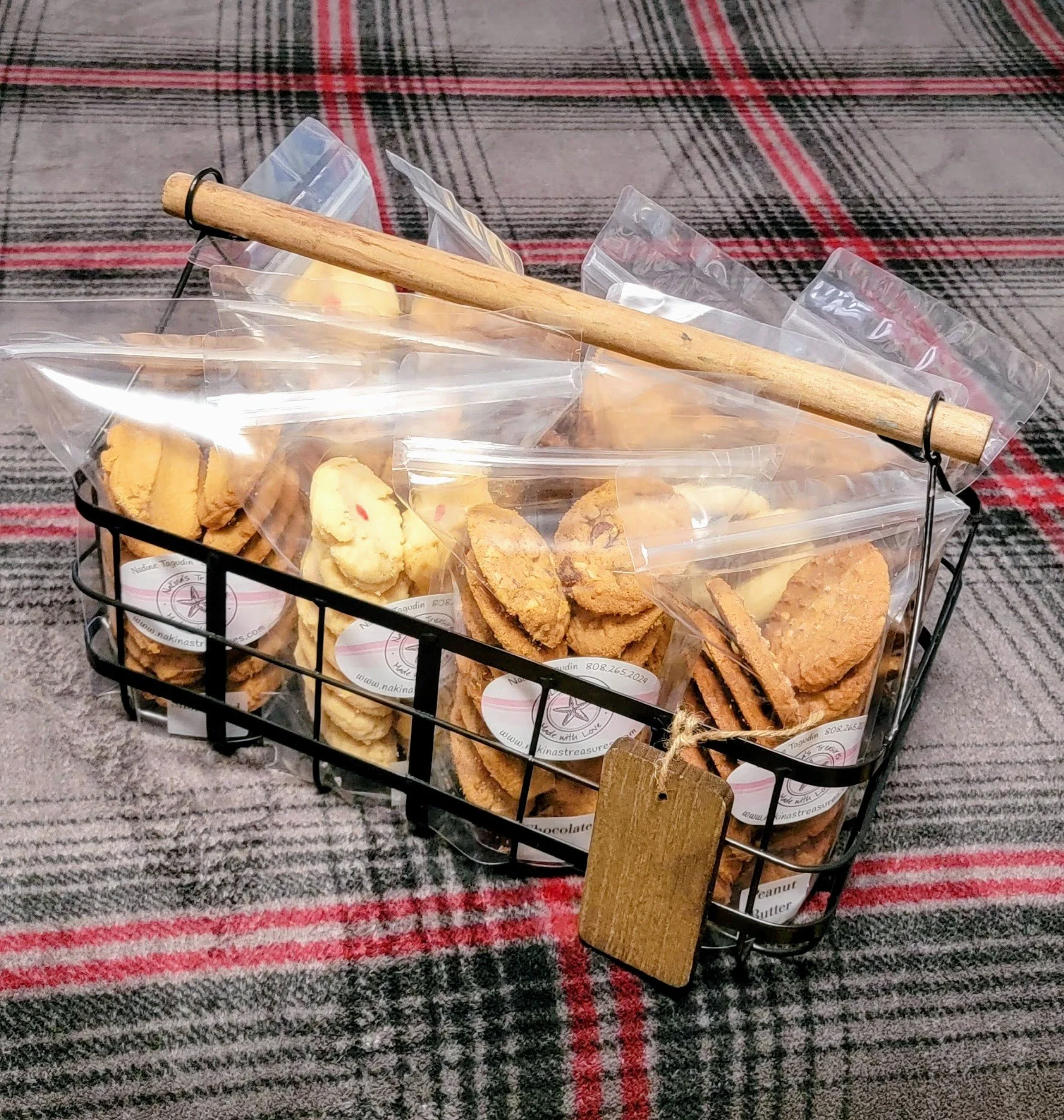 A black metal basket filled with various packaged cookies, placed on a red, black, and gray plaid fabric surface, with a small wooden board and a baking bundle tied with a string attached to the basket.