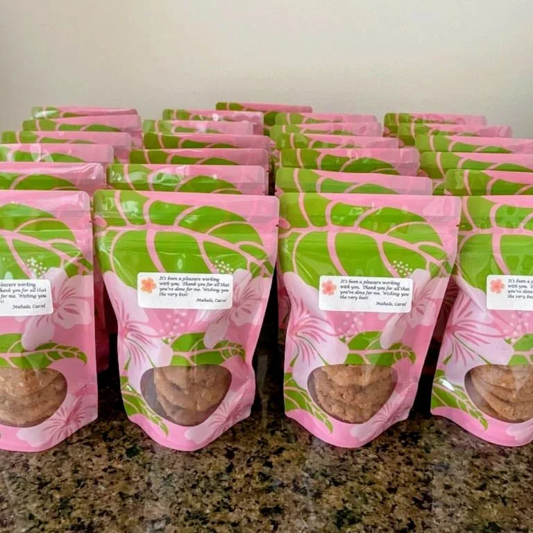 Multiple pink and green floral patterned snack bags lined up on a granite countertop, each with a clear window showing cookies inside and a small sticker with a thank you message.