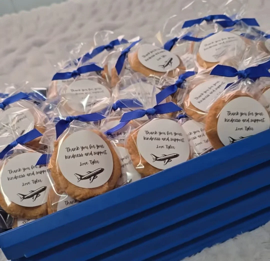 A blue tray filled with individually wrapped cookies, each with a white circular label featuring an airplane illustration and a message of thanks for kindness and support, signed by Tyler.