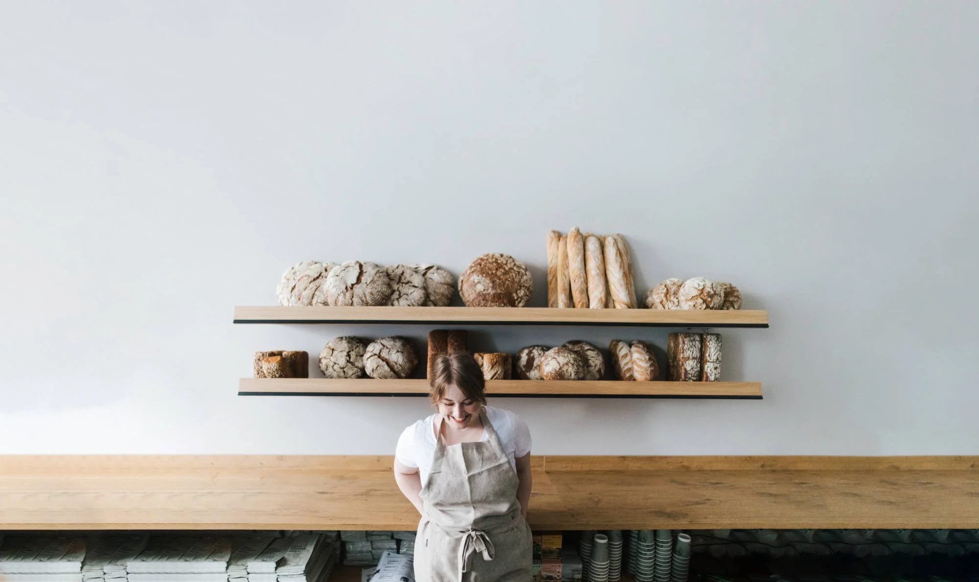 Eine junge Frau trägt eine Schürze und lächelt, während sie hinter einem Tresen in einer Bäckerei steht. Hinter ihr sind zwei Holzregale mit verschiedenen Broten und Gebäck, die auf den Regalen platziert sind.