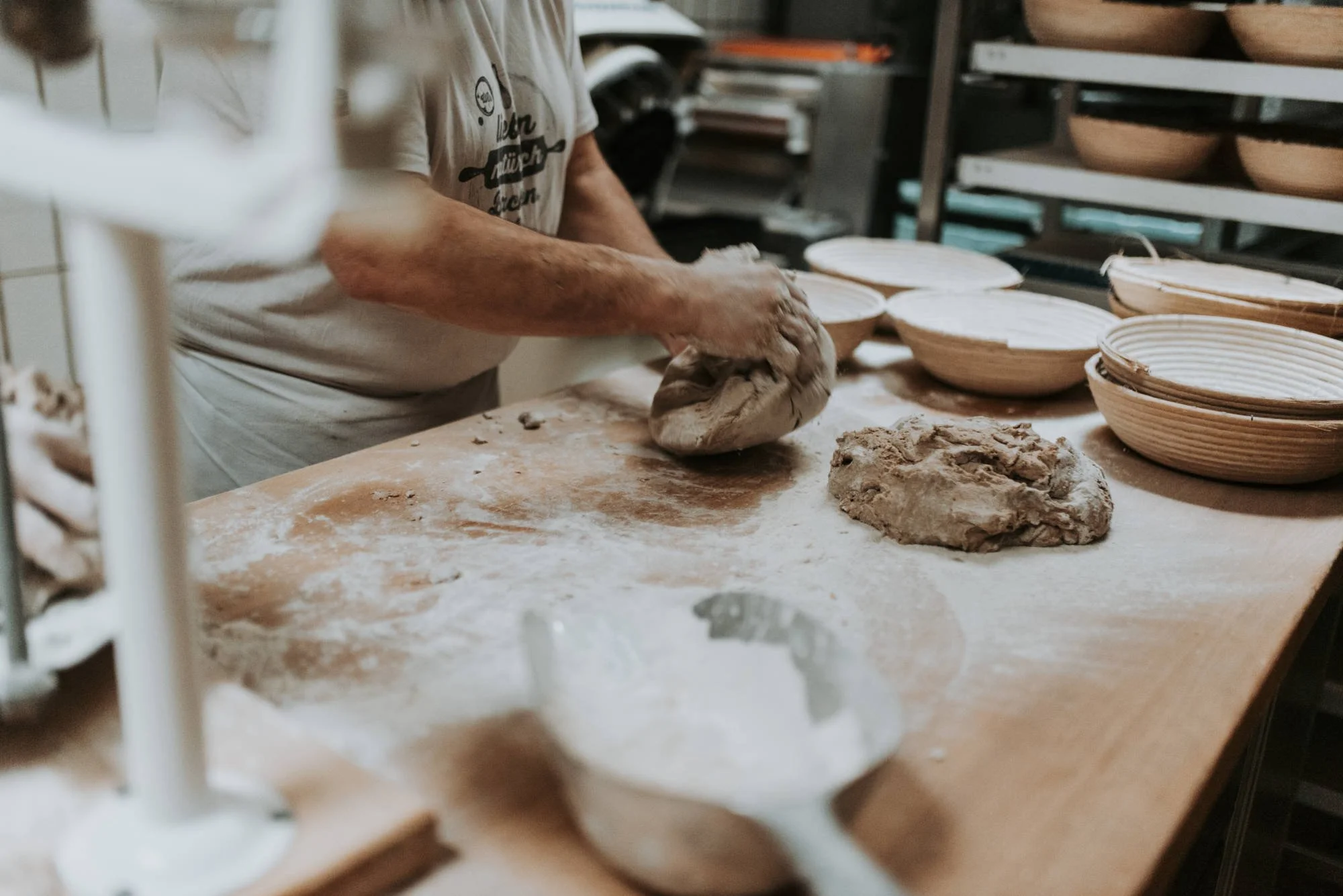 Eine Person arbeitet in einer Werkstatt an keramischen Schüsseln, mit Ton auf einem Arbeitstisch. Im Hintergrund sind mehrere Regale mit kleinen Holzschüsseln.