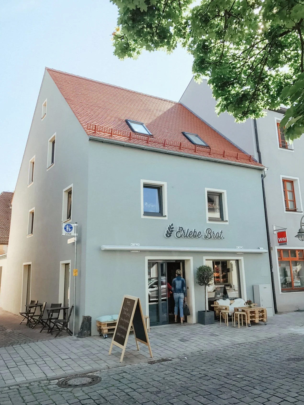 Ein dreigeschossiges, hellblau gestrichenes Gebäude mit roten Ziegeldächern. Es hat eine Bäckerei namens 'Erlebe Brot', erkennbar an einem Schriftzug und einem Brotlogo. Vor dem Laden stehen Tische und Stühle, und eine Person betritt den Laden.