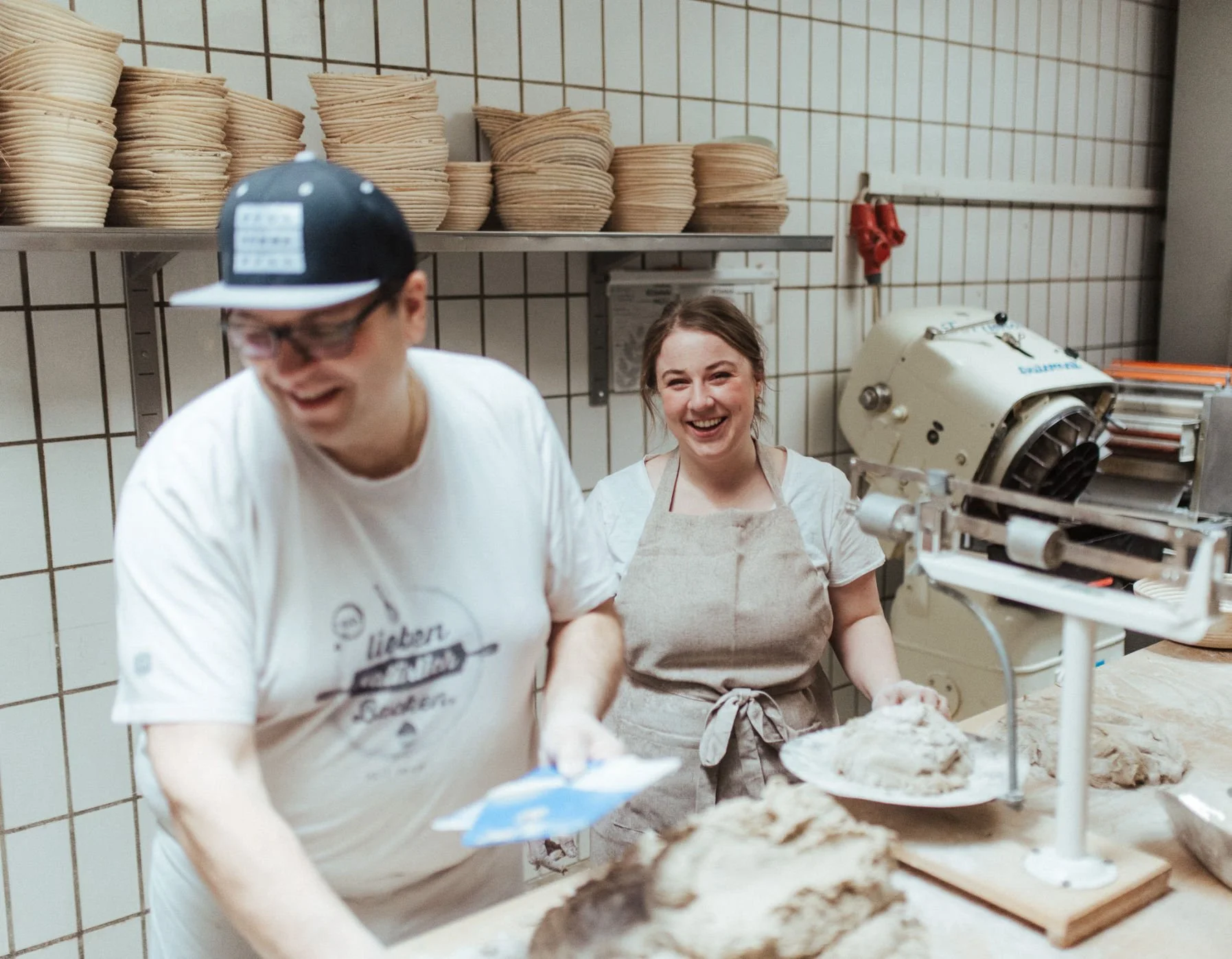 Zwei lachende Frauen beim Teig machen in einer Bäckerei, mit einer Maschine und Stapeln von Holzschalen im Hintergrund.