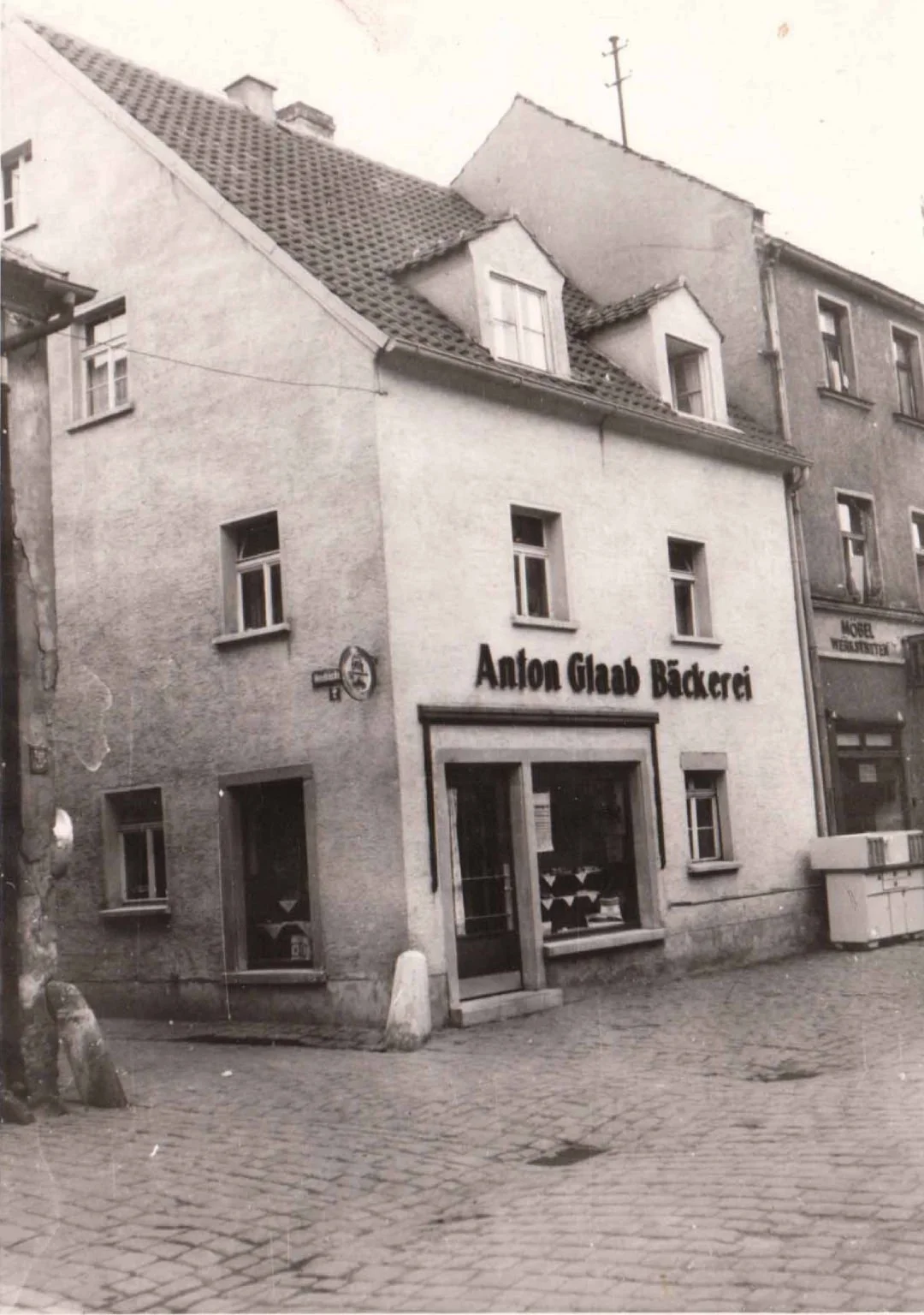Ein altes schwarzes und weißes Foto eines Gebäudes mit der Beschriftung 'Anton Glaab Bäckerei', das sich an einer gepflasterten Straße befindet. Das Gebäude hat mehrere Fenster, und es ist ein Laden mit großen Fenstern im Erdgeschoss.