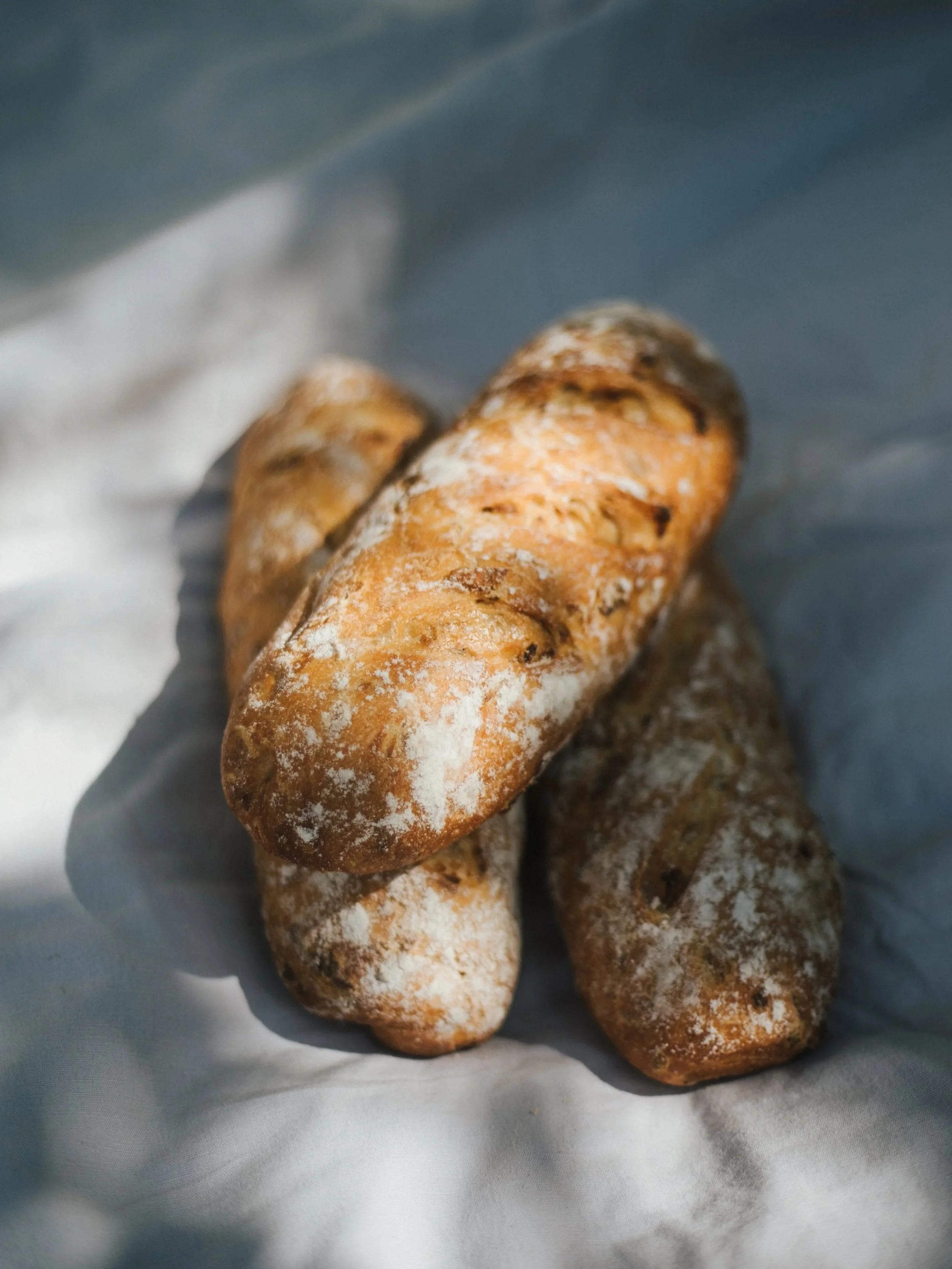 Mehrere Brote mit einer rustikalen, körnigen Kruste, bestäubt mit Mehl, liegen auf einem weißen Tuch.