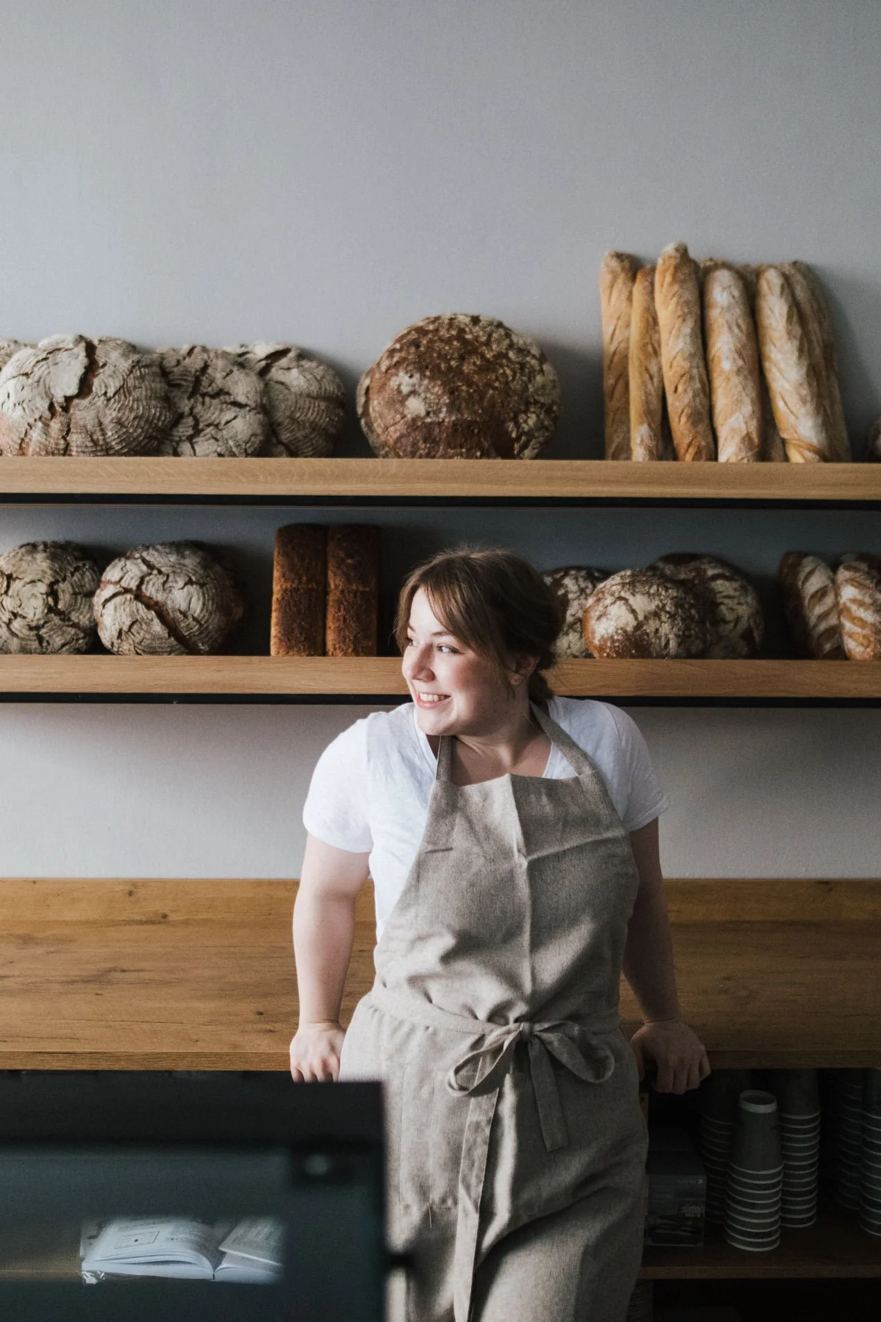 Eine lächelnde Frau mit braunen Haaren, trägt ein weißes T-Shirt und eine grau-gestreifte Schürze, steht in einer Bäckerei vor einer Holztheke, umgeben von Broten auf hohen Regalen im Hintergrund.